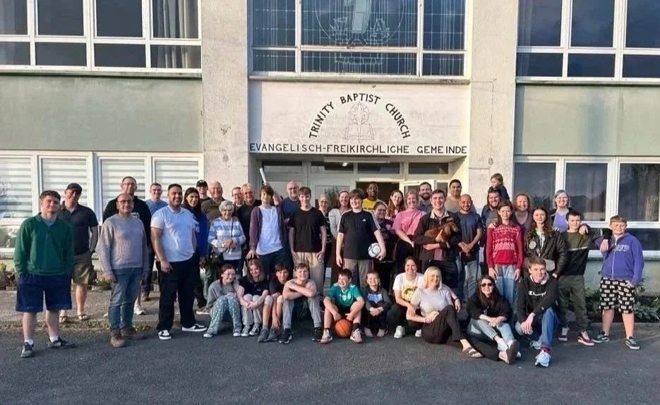 A group of people, including children and adults, standing outside the Trinity Baptist Church in front of the building entrance.