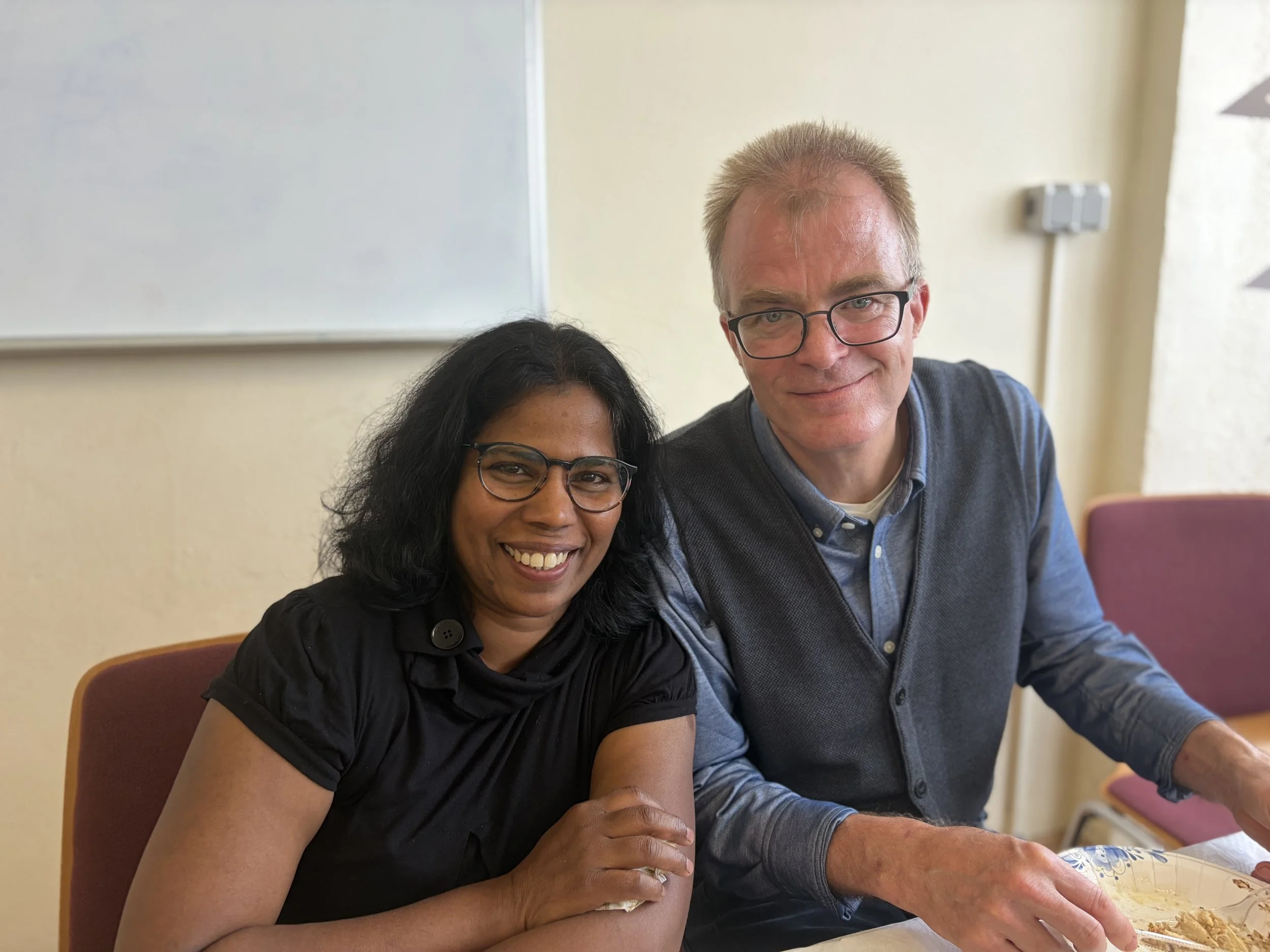 Smiling woman with dark hair and glasses sitting next to a smiling man with glasses, in a room with yellow walls and a whiteboard. They are sitting at a table with a floral tablecloth.