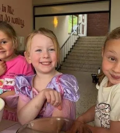 Three young girls smiling and sitting at a table inside a building with stairs in the background.