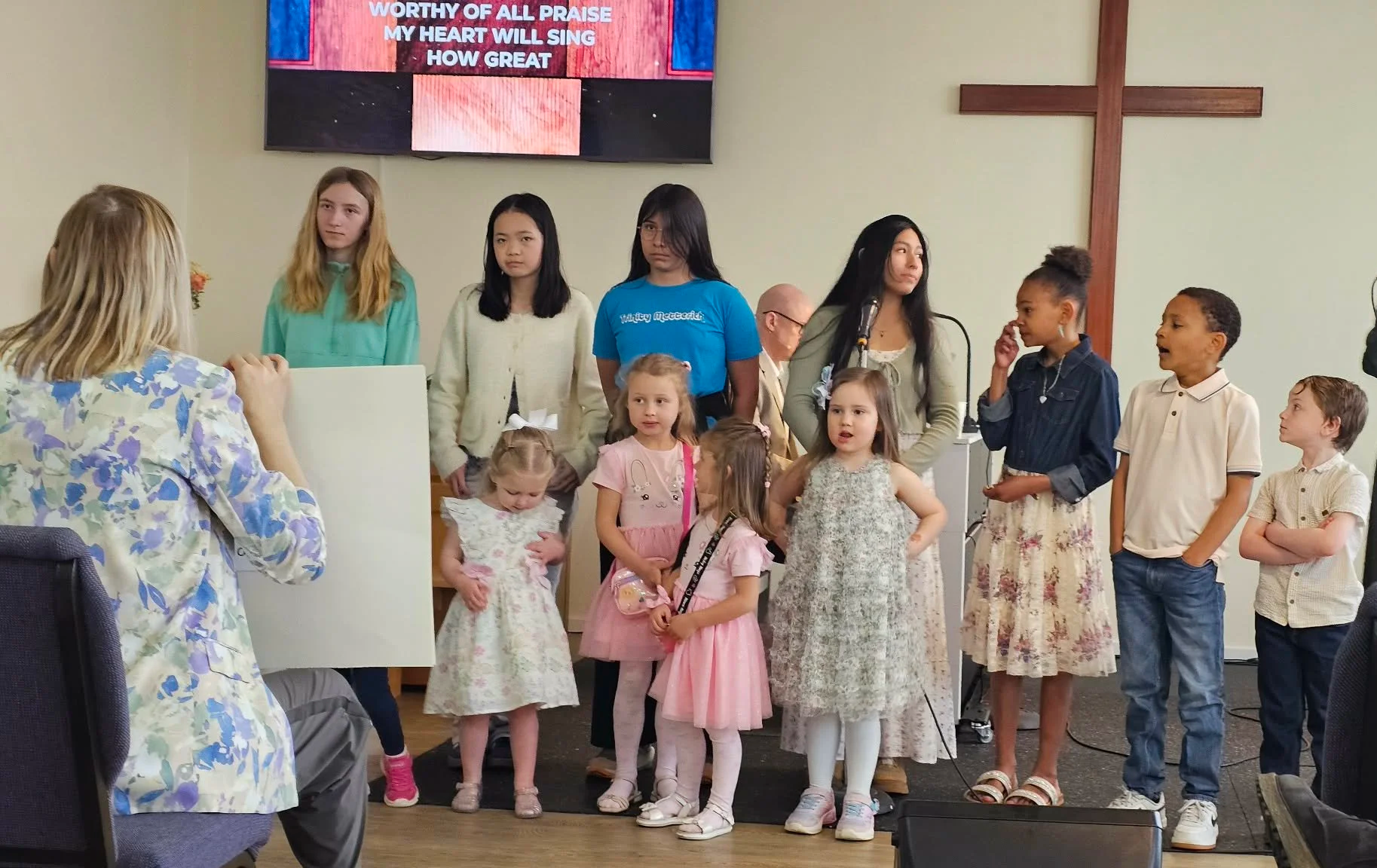 Children and adults gathered in a church for a hymn or praise service, with a cross on the wall and a large screen displaying lyrics.