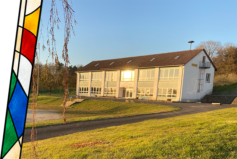 A white two-story building with large windows, set on a grassy hill during sunset, with a path leading up to the entrance and surrounding trees.