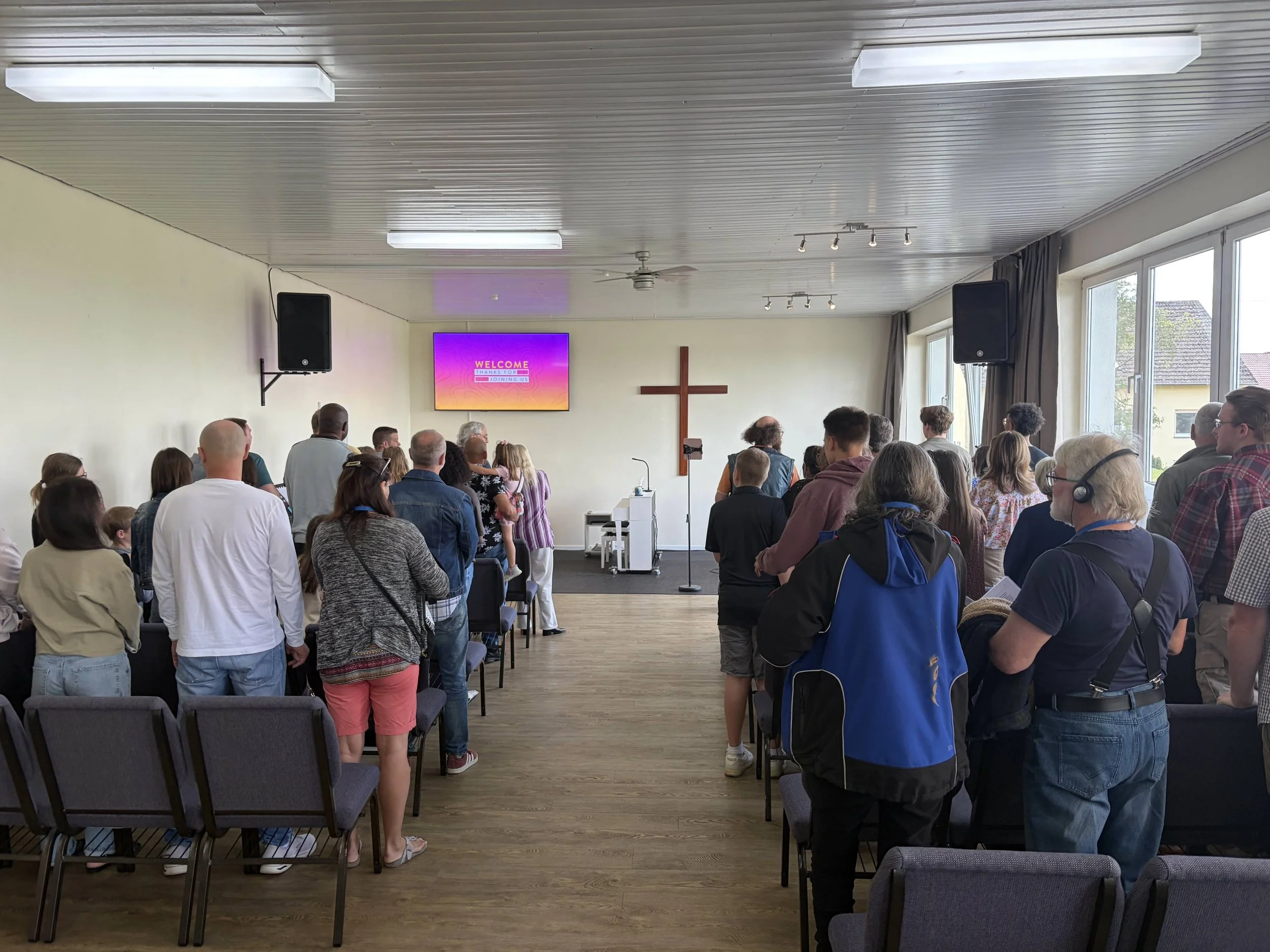People gathered inside a church or community hall, facing a stage with a large screen displaying a welcome message and a cross on the wall behind. Windows along the side wall let in natural light.
