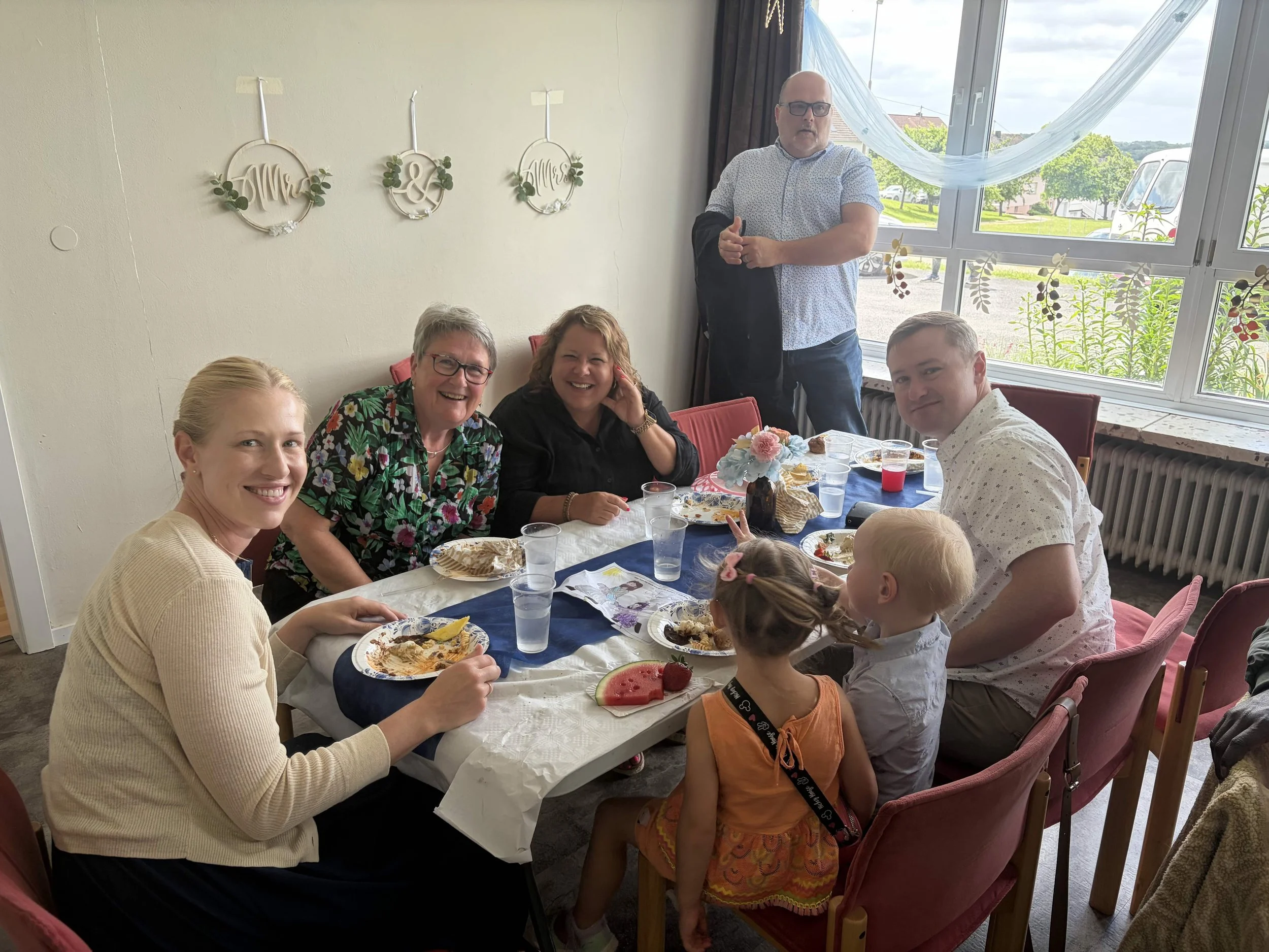 A group of seven people gathered around a dining table, enjoying a meal, with some children present. The setting is a bright room with large windows, floral decorations on the wall, and a table set with plates, drinks, and a centerpiece.