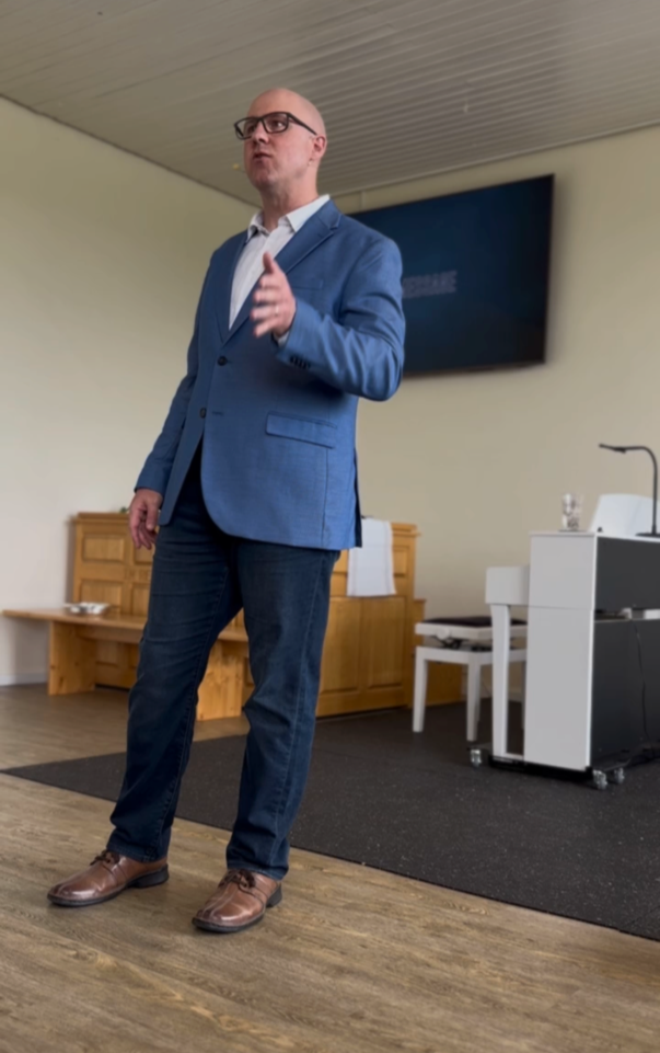 A man in a blue blazer, white shirt, and dark jeans standing in a room, gesturing with his right hand while speaking. There is a large TV screen behind him, a white desk with a microphone, and a wooden bench on the wall.