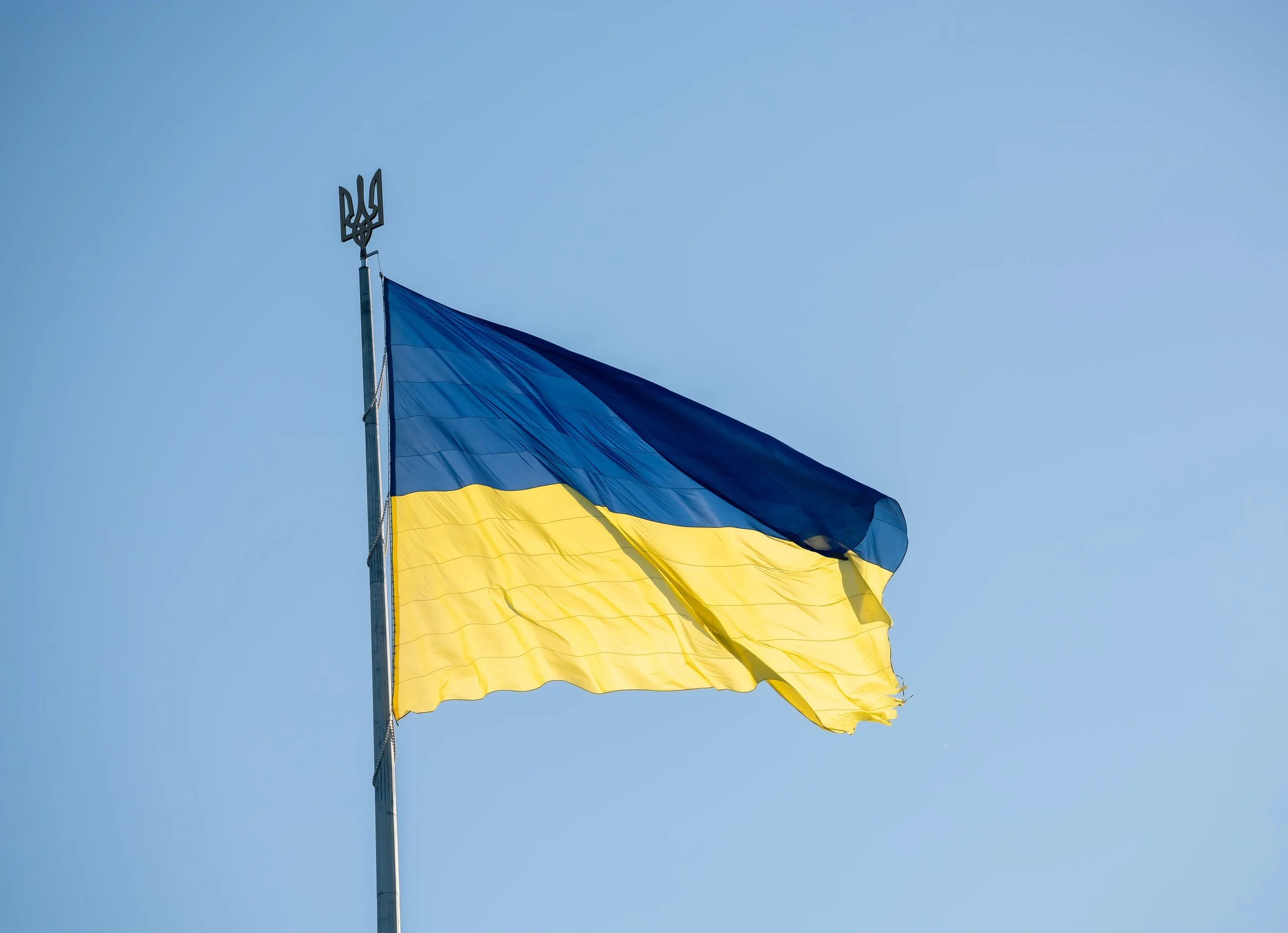 Ukrainian flag waving in the wind against a clear blue sky, with a small emblem of Ukraine's coat of arms at the top of the flagpole.