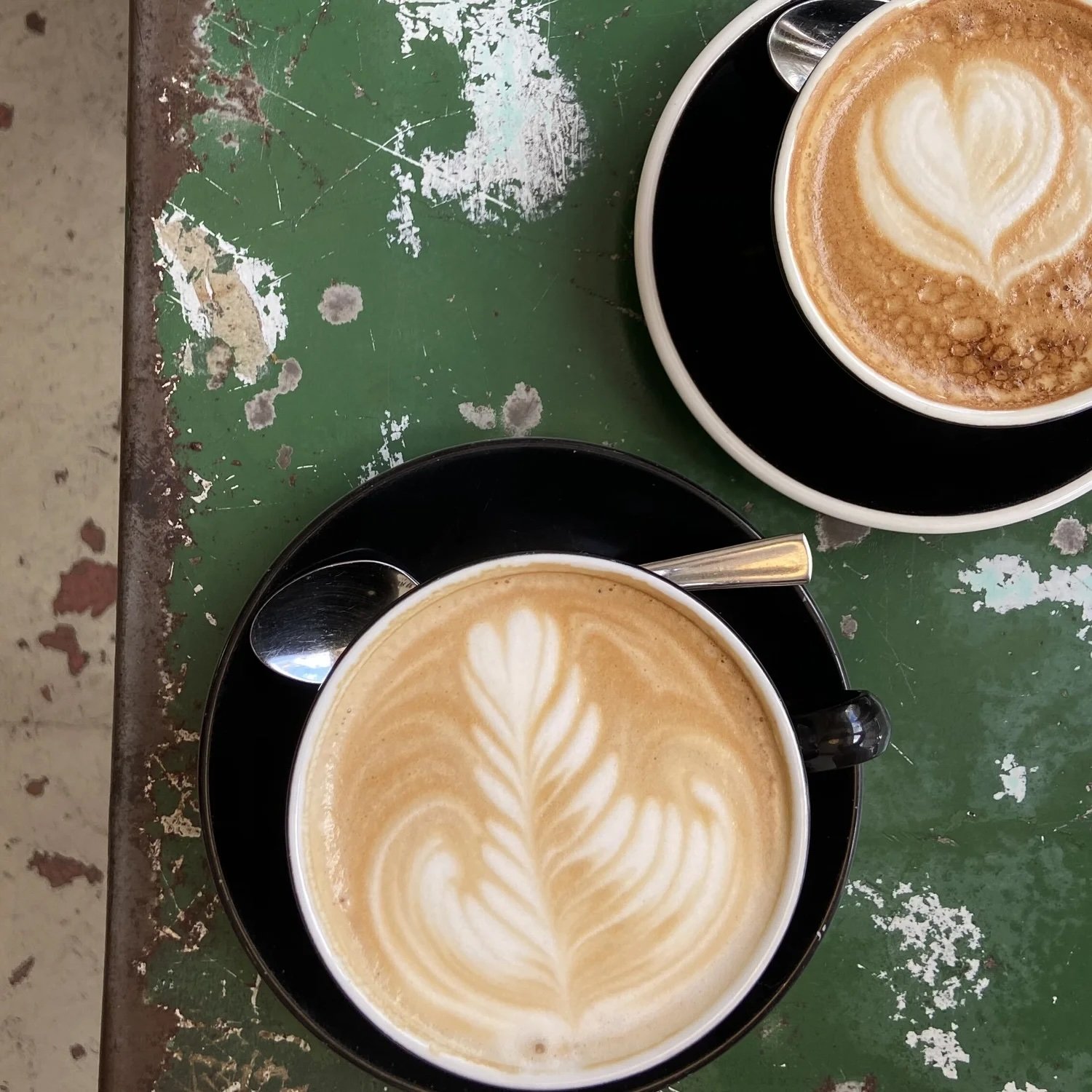 Two cups of latte with latte art on a green, weathered table. Each cup has a spoon resting on the saucer.