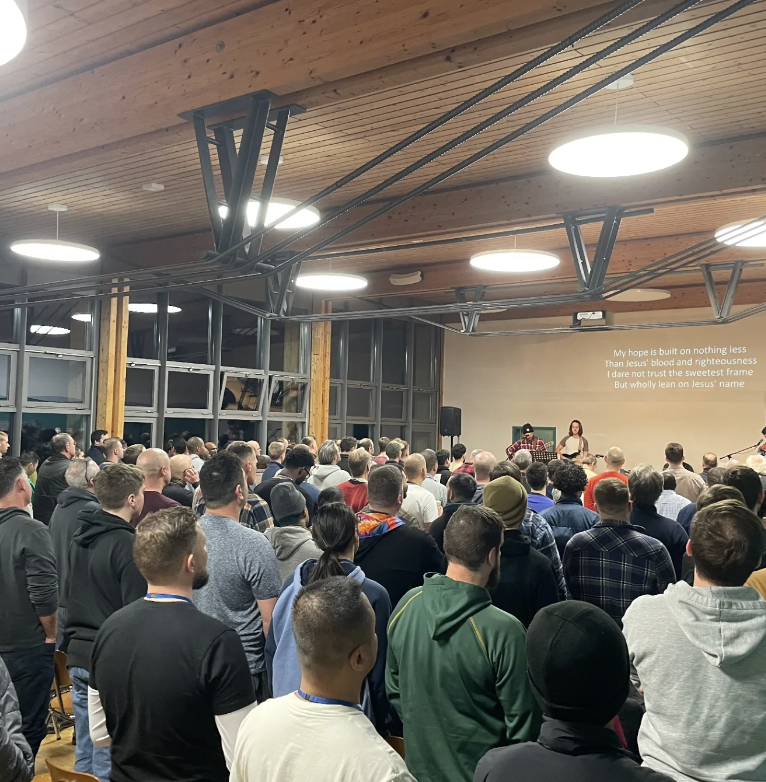 Crowd attending a musical performance in a wooden ceiling venue, with lyrics projected on the wall.
