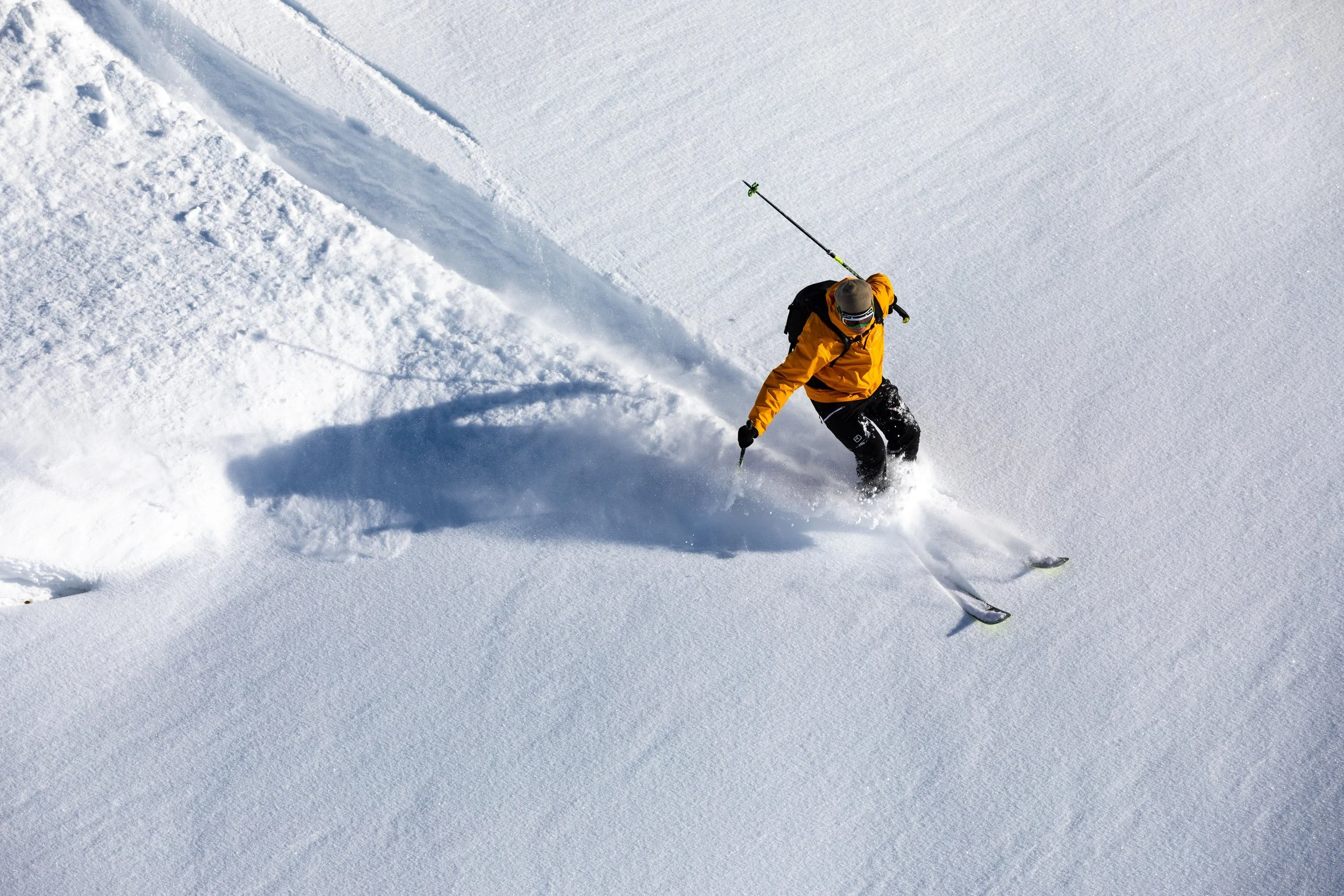 A person wearing an orange jacket, black pants, and a gray hat skiing in deep snow, creating a trail behind them.
