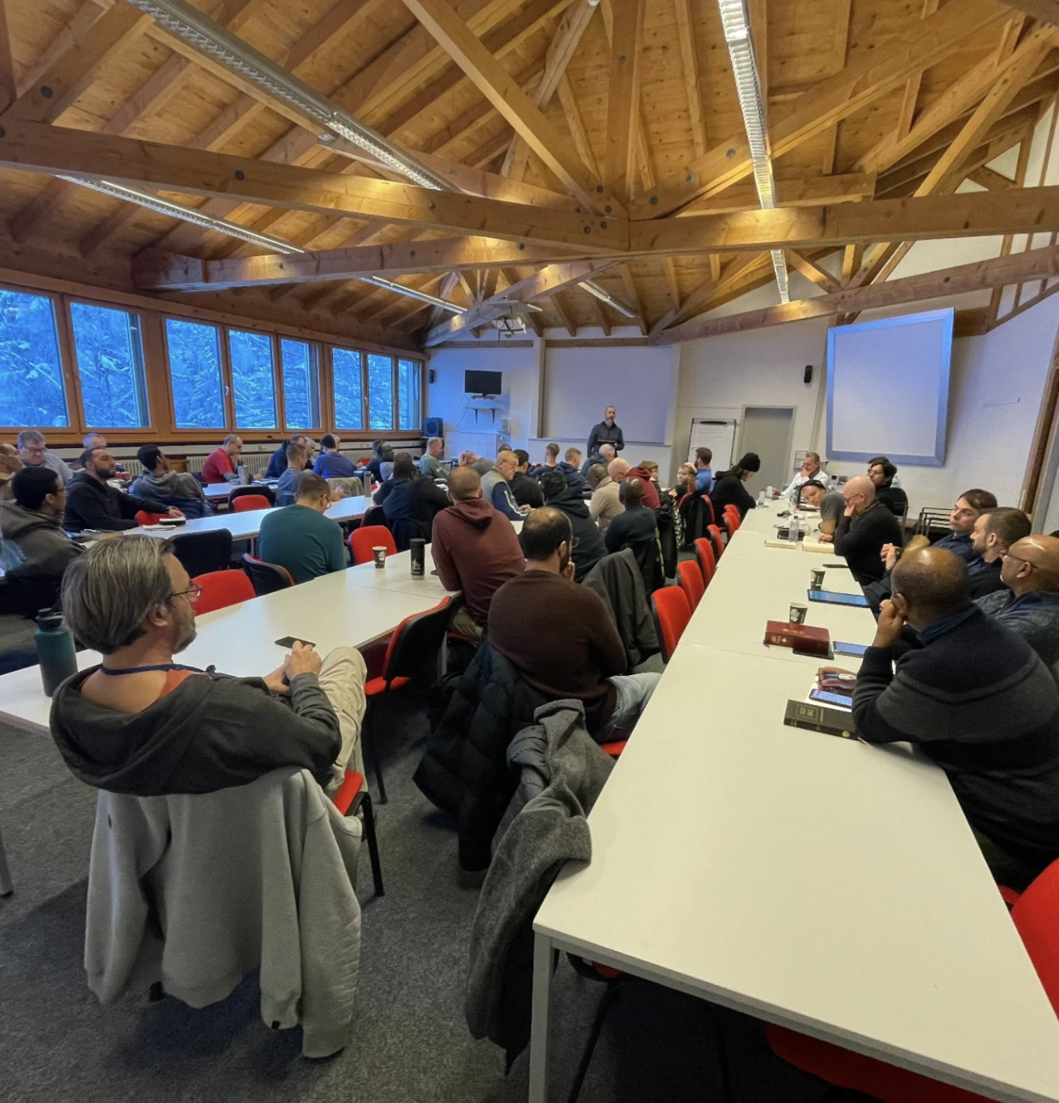 A group of people attending a conference or workshop in a large room with wooden ceiling beams, large windows showing a snowy landscape outside, some people taking notes, and a speaker standing at the front.