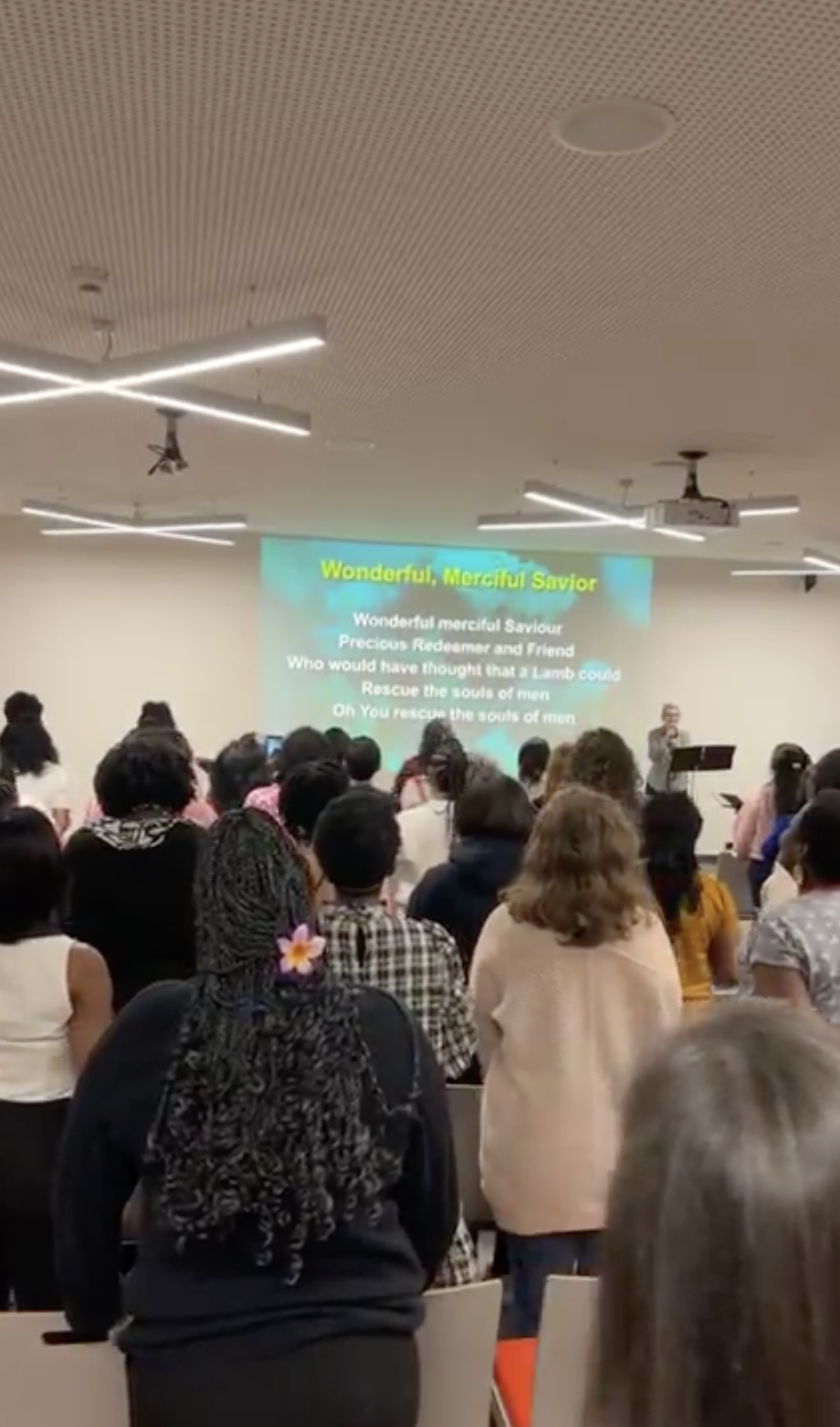 A crowded room with people attending a worship service or concert, facing a stage with a large screen displaying song lyrics titled "Wonderful, Merciful Savior." The screen shows lyrics about a savior who rescues souls. The room has modern lighting and ceiling fixtures.