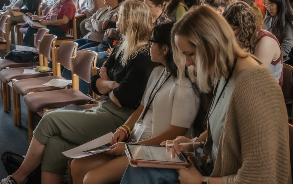 People sitting in rows at a conference or seminar, some using tablets or reading papers.