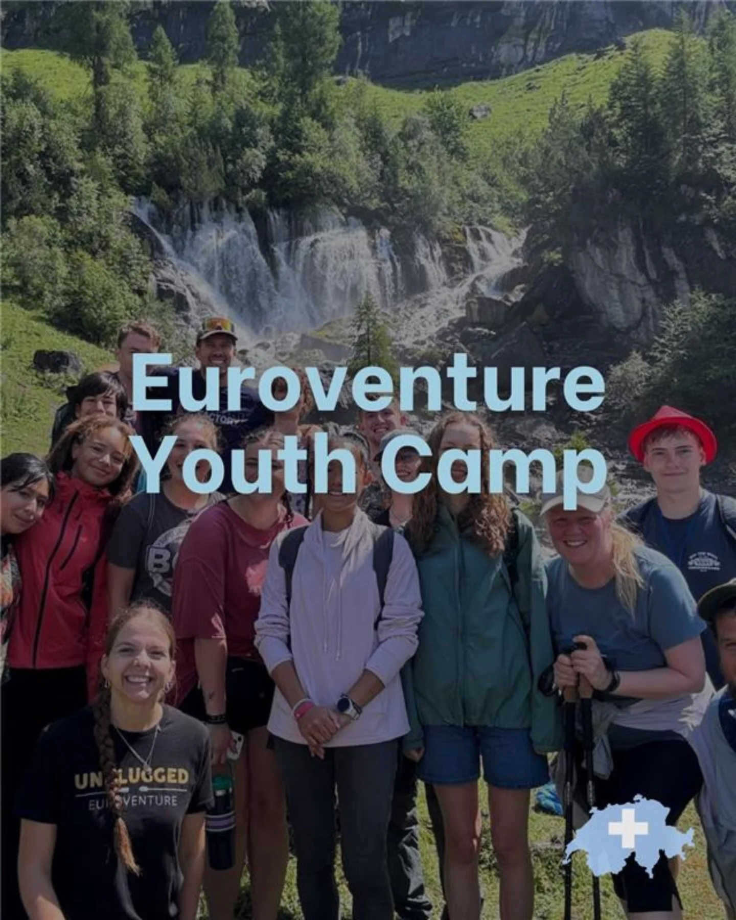 Group of young people smiling and standing together outdoors in front of a waterfall and lush green landscape with mountains on a sunny day. The photo features diverse individuals, some wearing backpacks and outdoor gear, and a few holding sunglasses.