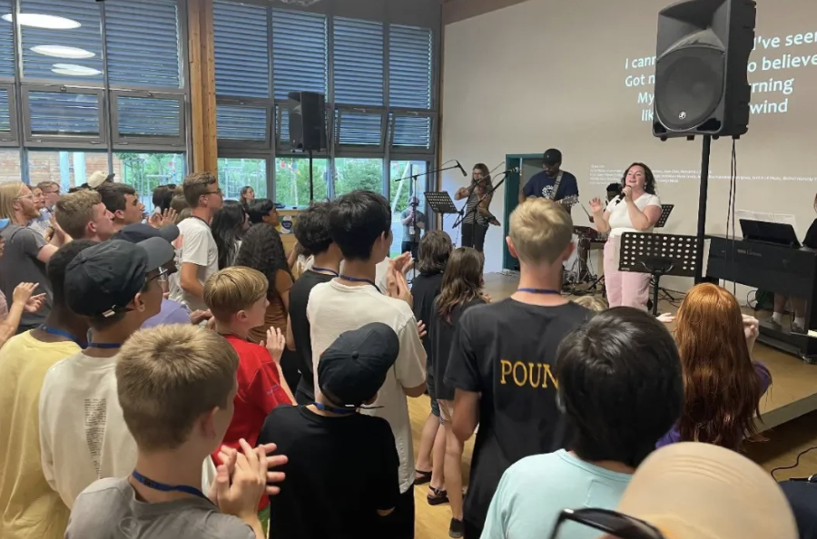 A diverse group of young people attending a church service or concert, with some dancing and singing along to a live band on stage, which includes a female singer, guitarists, and a violinist in a room with large windows and a screen displaying lyrics.