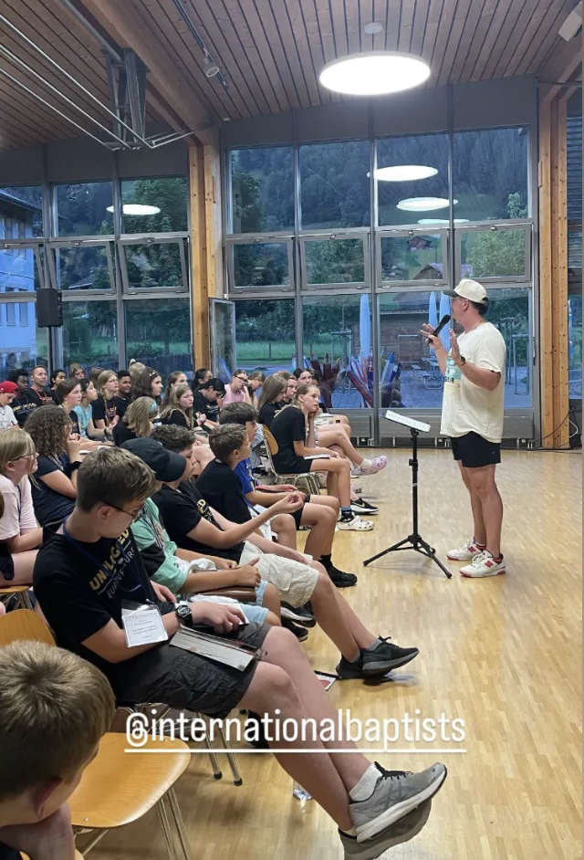 A group of children and teenagers seated in a large room, listening to a speaker standing in front of them. The speaker is wearing a beige cap, a light-colored t-shirt, and shorts, and is looking at their phone. The room has large windows, wooden walls and ceiling, and circular ceiling lights.