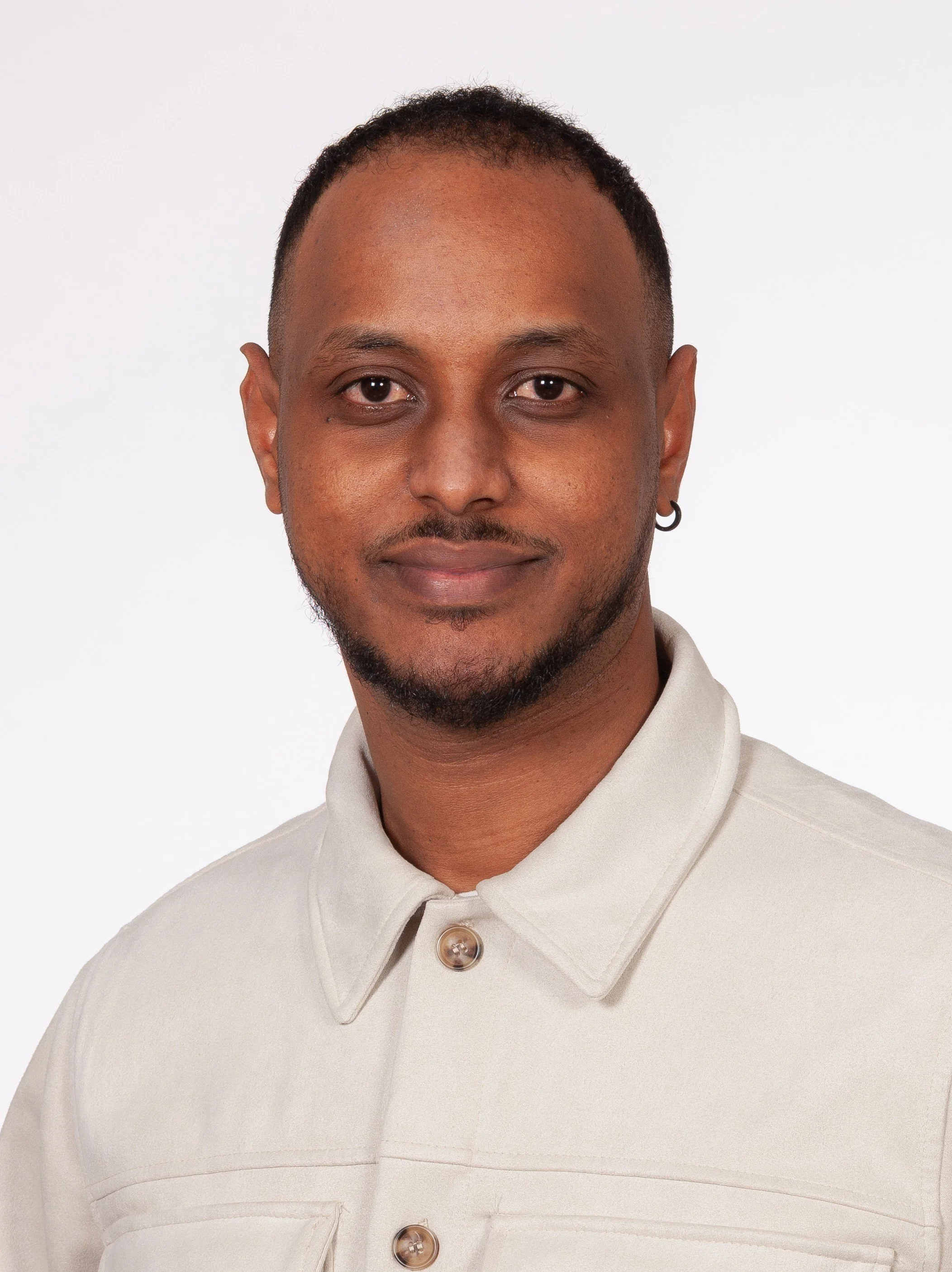 Ein Mann mit dunkler Haut, kurzen schwarzen Haaren, Träne im Ohr, trägt einen beige Hemd mit Knöpfen, lächelt leicht, sitzt vor weißem Hintergrund.