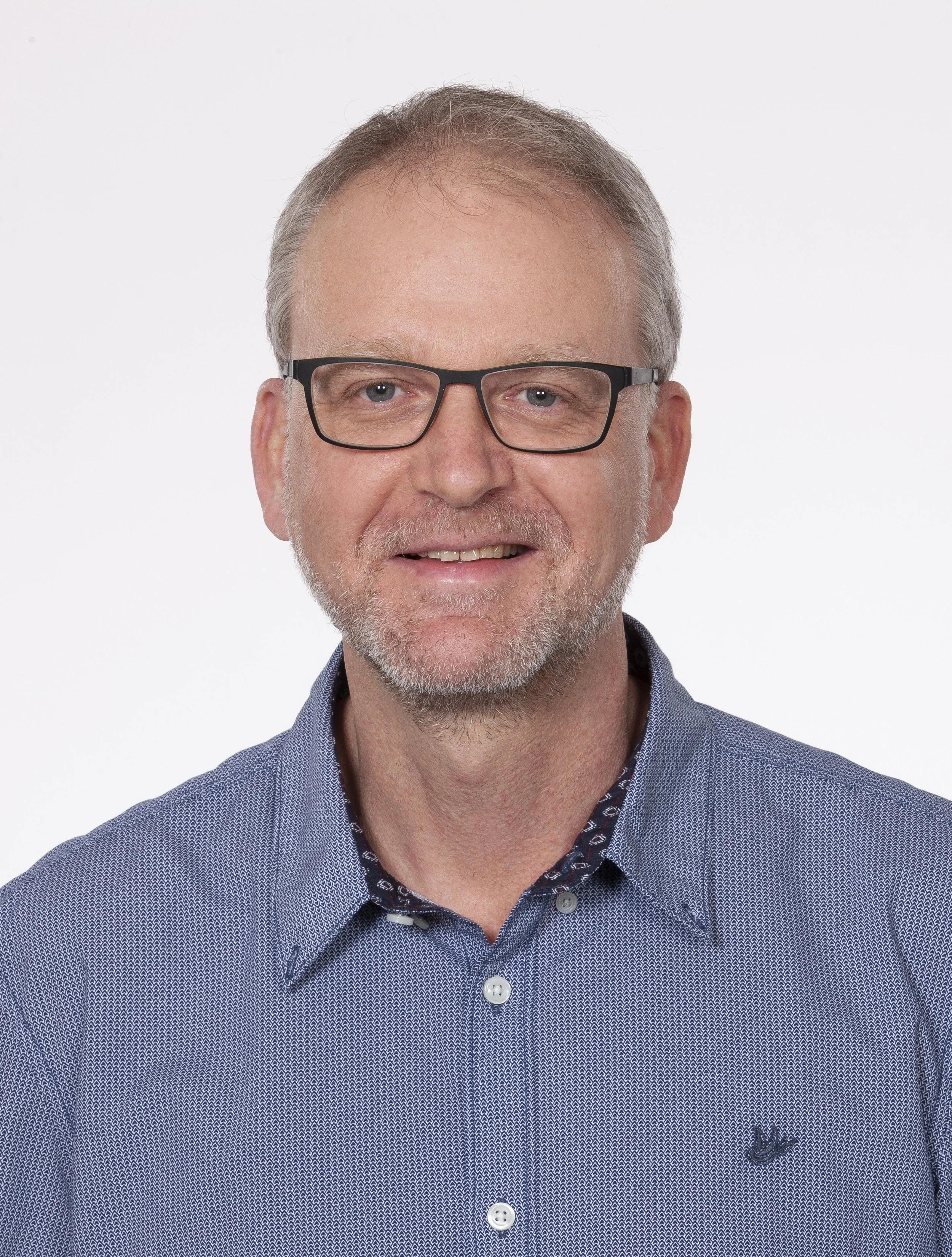 Ein älterer Mann mit Brille, grauem Haar und Bart trägt ein blaues Hemd mit einem kleinen Logo auf der Brust, lächelt vor einem weißen Hintergrund.