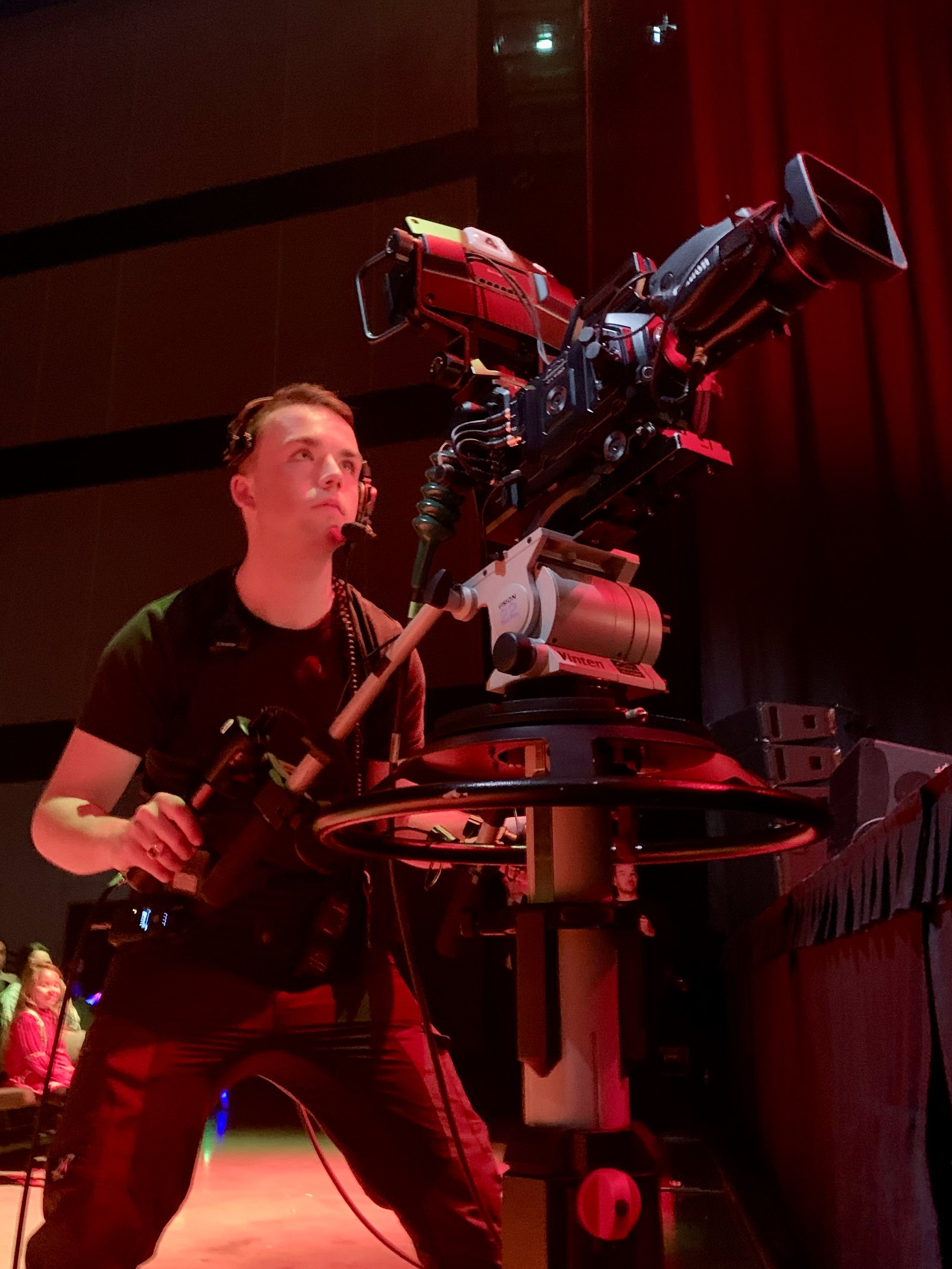 Cameraman met headset bedient een professionele videocamera op een podium tijdens een evenement.