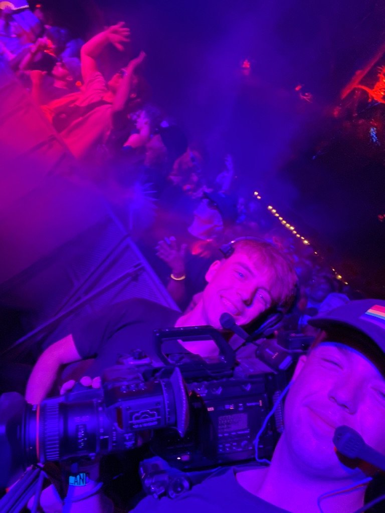 Twee personen nemen een selfie met een grote videocamera bij een concert of festival, omgeven door paarse en roze verlichting, terwijl mensen op de achtergrond dansen.