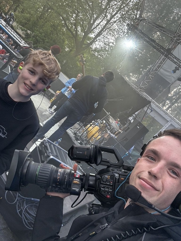 Twee mensen op een muziekpodium, een met een camera en de ander met in-ear monitors. Op de achtergrond een band die speelt.