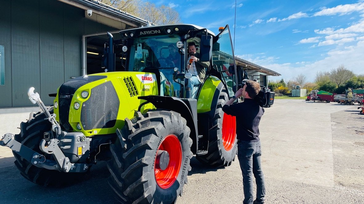 Man filmt een tractor Claas Arion 660 naast een loods op een zonnige dag.