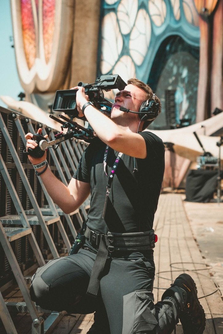 Een cameraman knielt op een podium met een professionele videocamera. Hij draagt een headset en houdt de camera omhoog gericht. Op de achtergrond zijn delen van decor en apparatuur zichtbaar.
