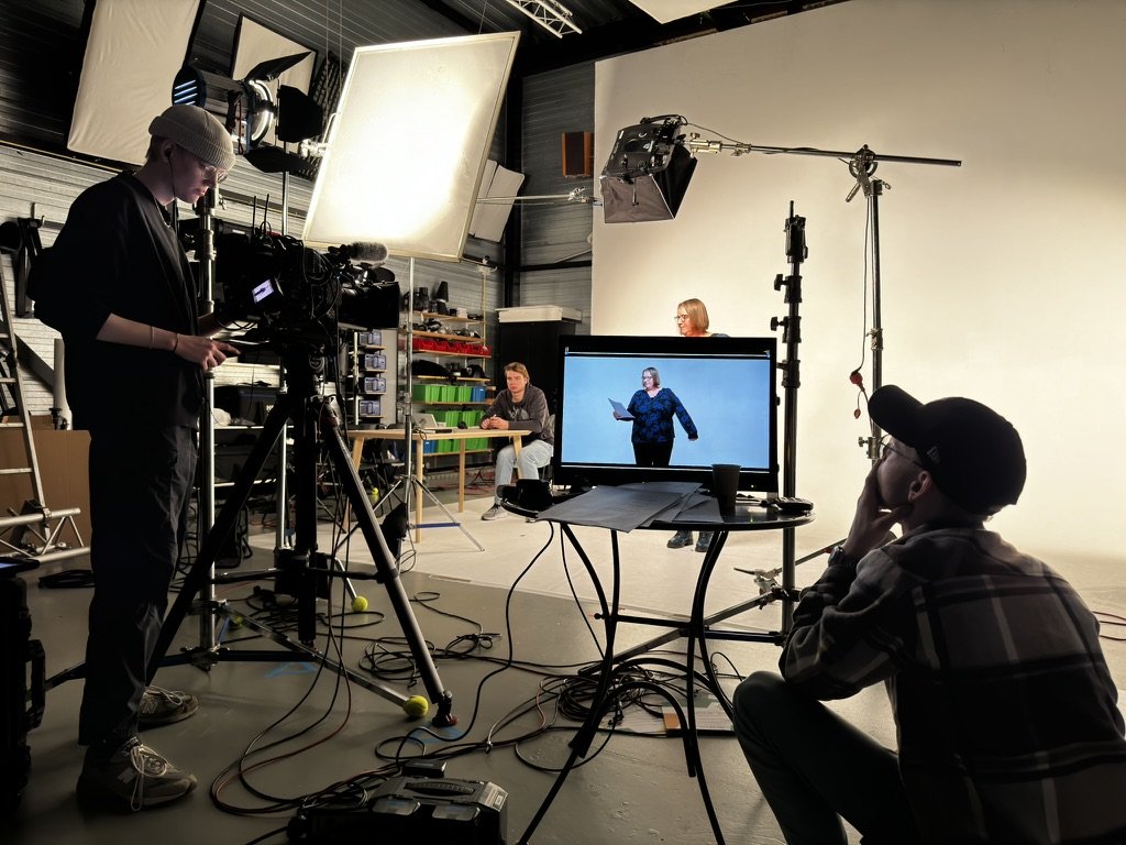 Opnamestudio met drie personen, camera-apparatuur, belichting, monitor die een vrouw laat zien met een tablet. Achtergrond is wit, studio heeft technische apparatuur en kabels verspreid over de vloer.