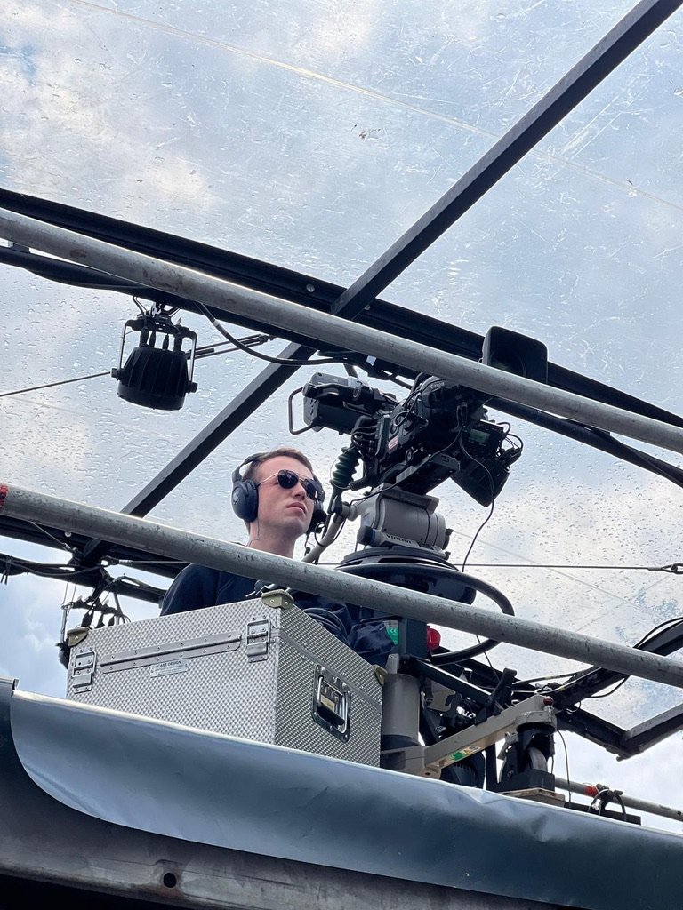 Persoon opereert een camera met koptelefoon en bril, omgeven door apparatuur, onder een glazen dak.