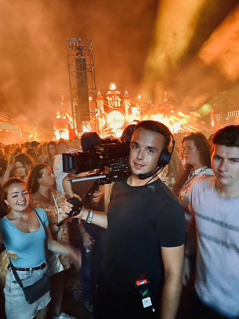 Een cameraman filmt tijdens een festival met lichtshows en een groot podium op de achtergrond. Er zijn veel mensen op de voorgrond.