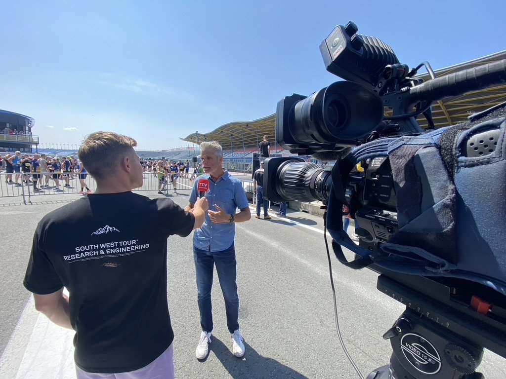 Een tv-interviewer op een racecircuit interviewt een man, terwijl een camera hen filmt. Op de achtergrond staan toeschouwers achter hekken. De lucht is helder en blauw.