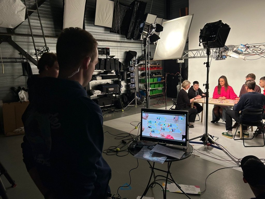 Een fotostudio met een groep mensen die rond een tafel zitten, bezig met een project. Er zijn studioverlichting, camerapersoneel en een monitor die de scène toont.