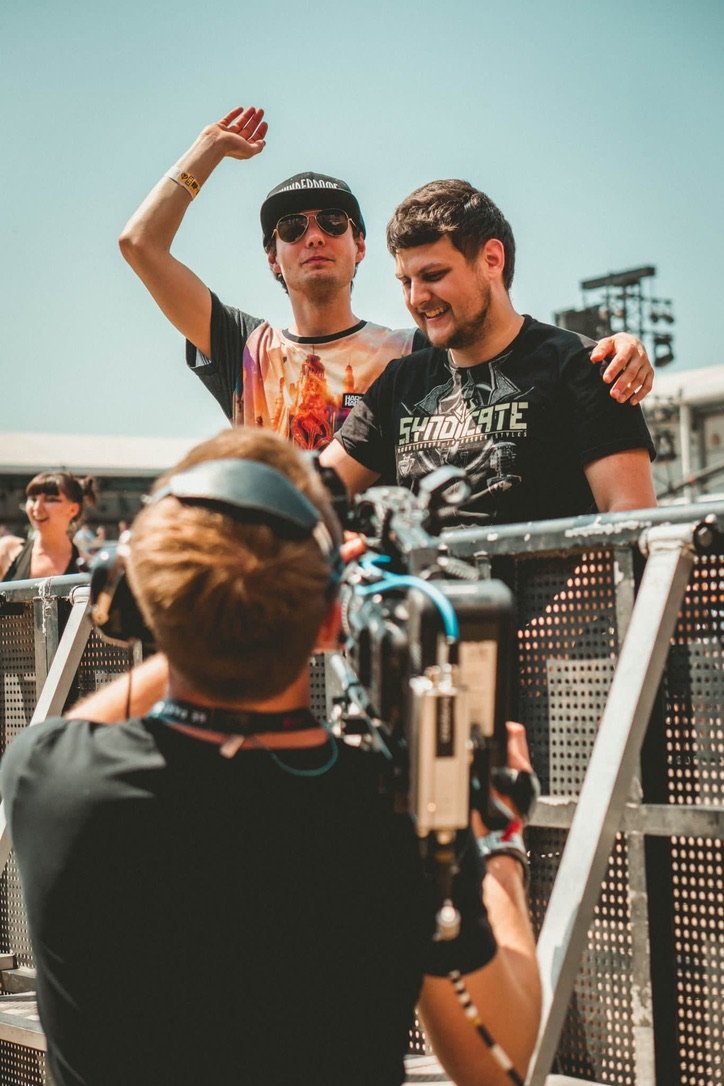 Twee mannen poseren voor een camera op een buitenlocatie. De man links draagt een pet en zonnebril, terwijl de man rechts lacht. Een cameraman filmt hen. Op de achtergrond zijn andere mensen zichtbaar.