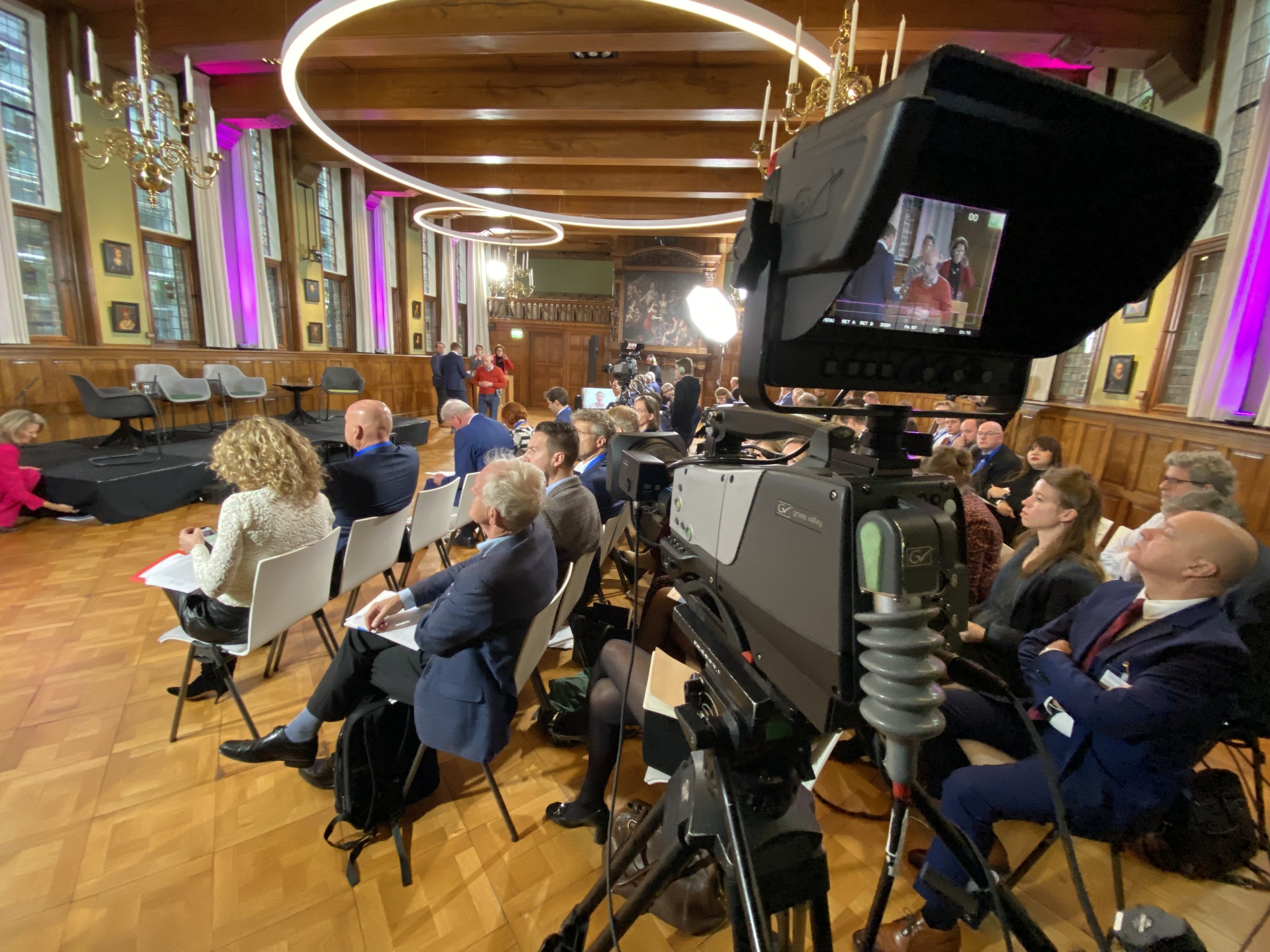 Conferentie of seminar in een ruime zaal met houten interieur, waar mensen op stoelen zitten gericht naar een podium met lege stoelen en microfoons. Een grote camera staat vooraan, en er zijn verlichte kroonluchters aan het plafond.