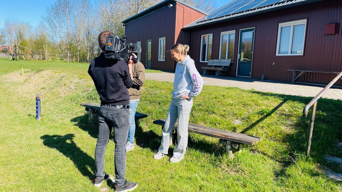 Twee mensen in gesprek buiten een gebouw, één persoon bedient een videocamera. Het gebouw heeft zonnepanelen en bevindt zich op een grasveld.
