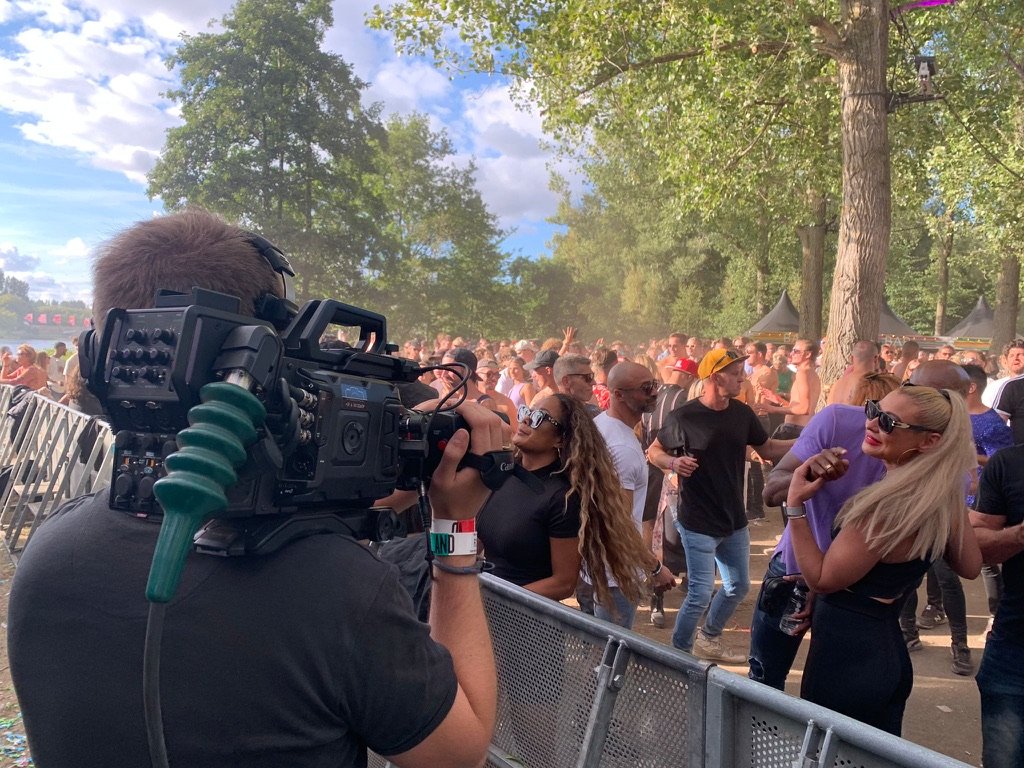 Mensen bij een buitenfestival; drukte met vele bezoekers en een cameraman in de voorgrond.