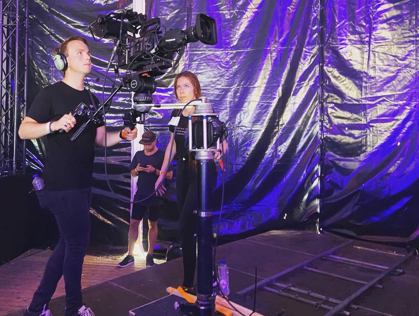 Twee mensen werken met een professionele filmcamera in een donkere omgeving met paarse verlichting. Een van hen heeft een koptelefoon op en bedient de camera op een dollyspoor, terwijl de ander naast hem staat. Op de achtergrond staat een derde perso