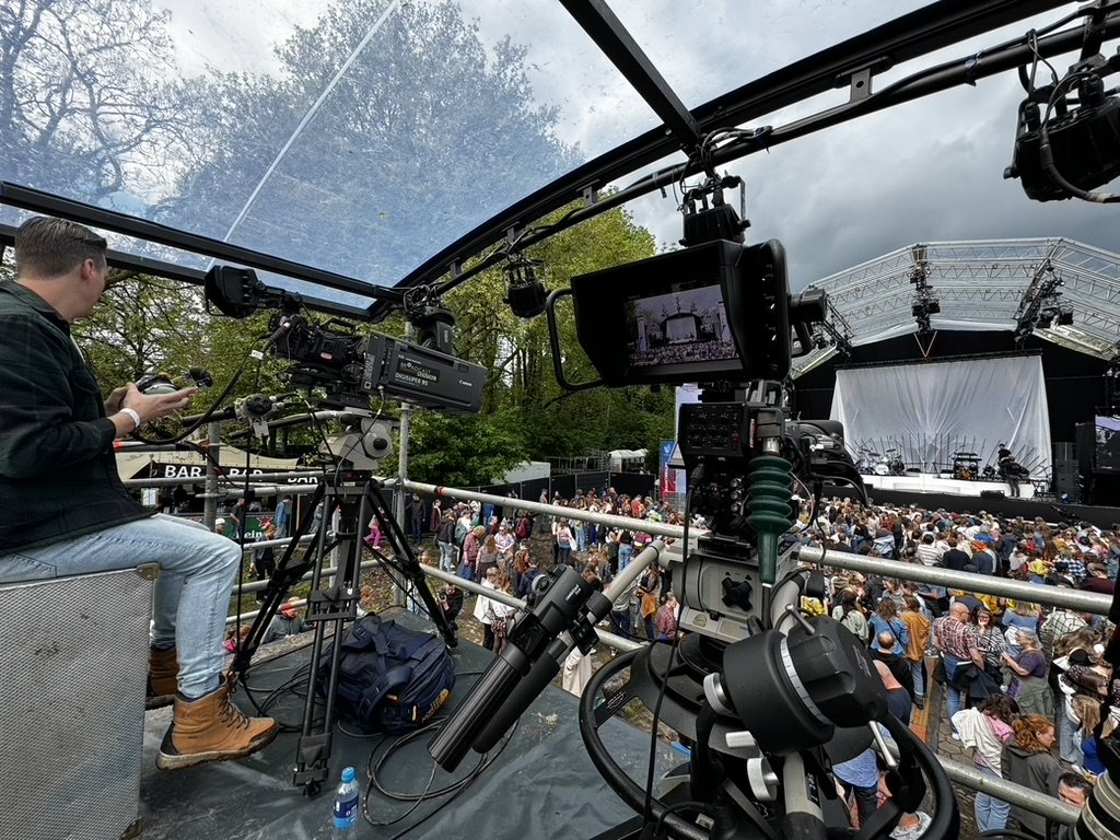 Grote scène met podium, een menigte mensen en professionele camera-apparatuur. Cameraman bedient apparatuur. Openluchtlocatie met overkapping en bomen in de achtergrond.
