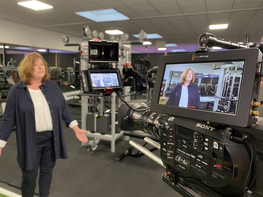 Een opname van een vrouw in een sportschool. Er staat een professionele camera voor haar, gericht op een beeldscherm dat de vrouw toont. De sportschool is goed verlicht, met fitnessapparatuur zichtbaar op de achtergrond.