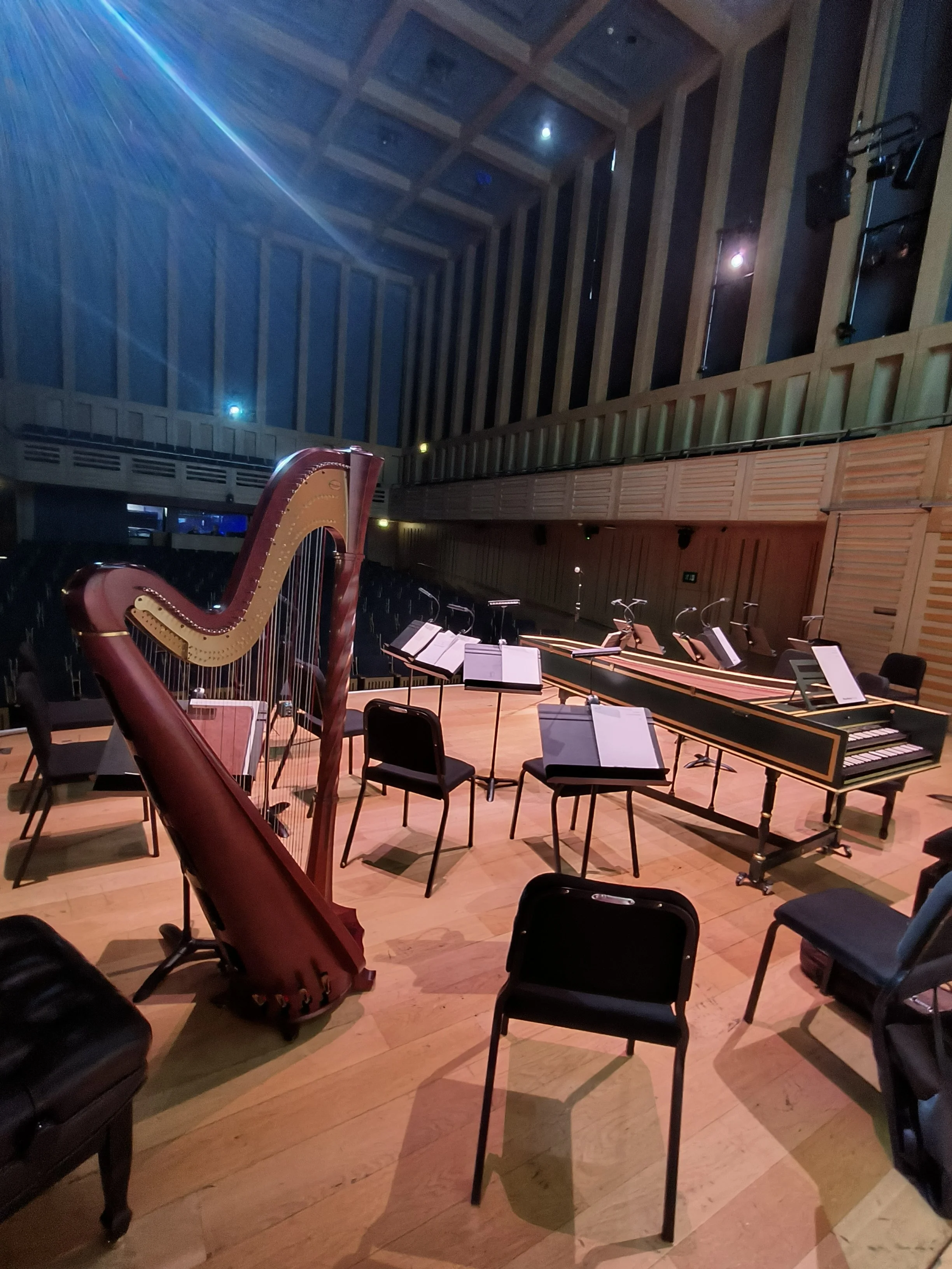 The concert hall at kings Place, London. Musical instruments including a harp, music stands, and chairs arranged for a performance. The hall features wooden flooring and slatted walls, with stage lights illuminating the space.