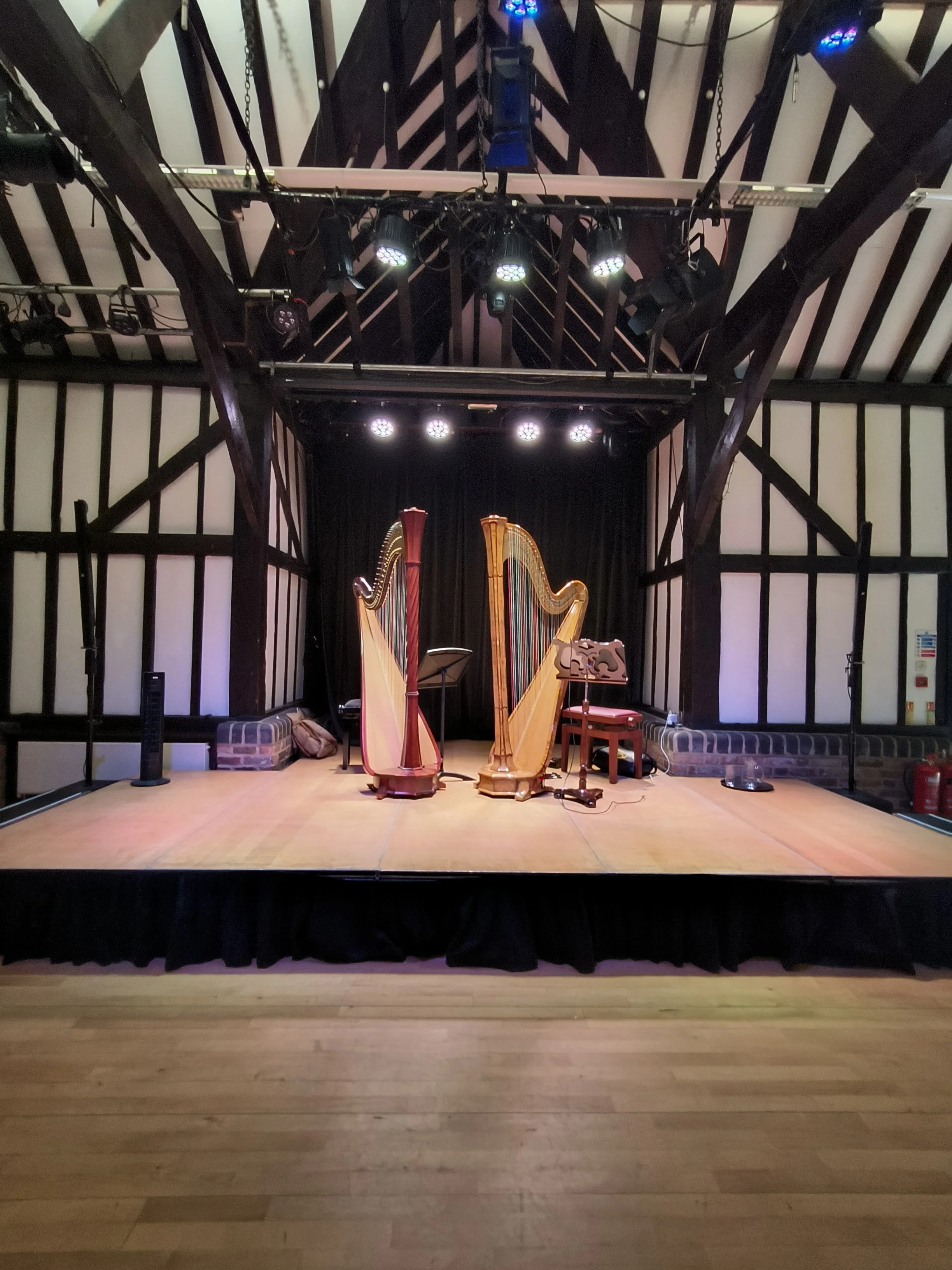 Stage with two harps, music stand, chair, and stage lights in a room with wooden floor and dark wooden beams on the ceiling.