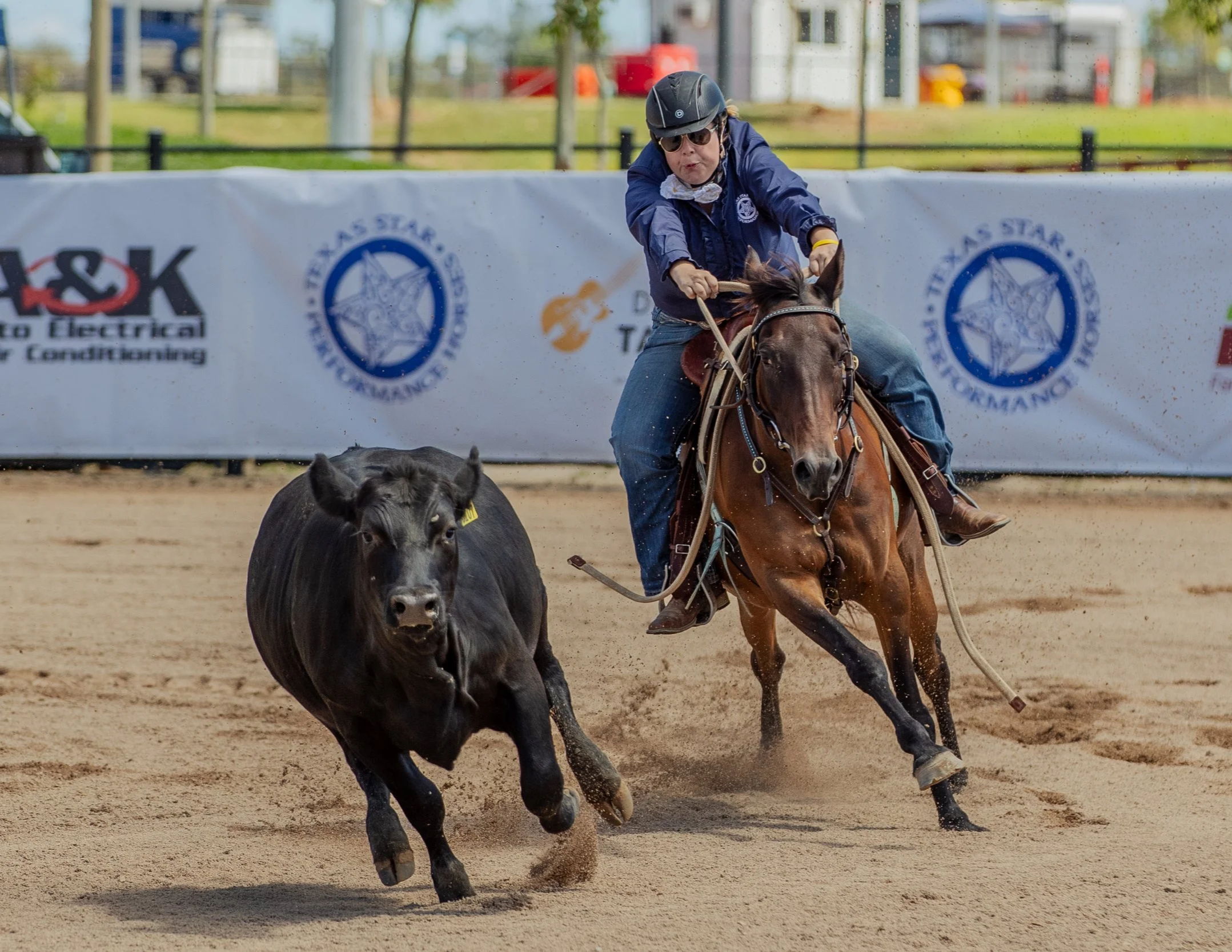 A rodeo competition showing a person on horseback chasing a cow on a dirt arena, with banners and structures in the background.