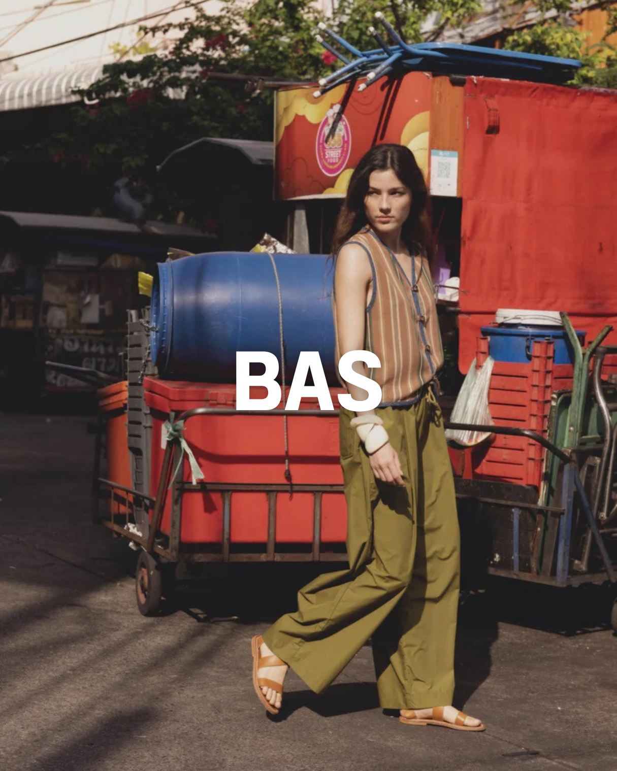 Une femme debout devant un chariot à marchandises, vêtue d'un pantalon ample olive, débardeur à rayures, sandales blanches et accessoires. En arrière-plan, un stand de rue avec des couleurs vives et un grand bidon bleu sur le chariot.