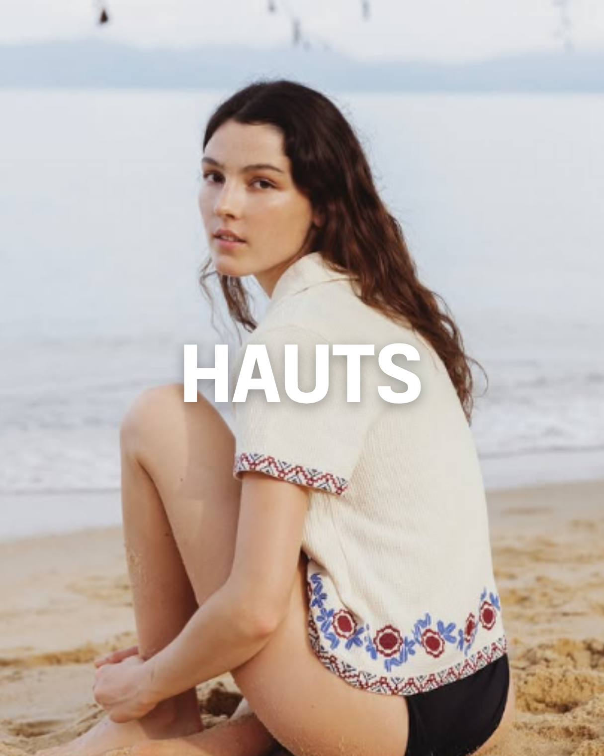 Jeune femme aux cheveux bruns, assise sur la plage, portant un haut d'été blanc avec des motifs colorés en rouge, bleu et noir, regardant vers la caméra, avec la mer en arrière-plan.