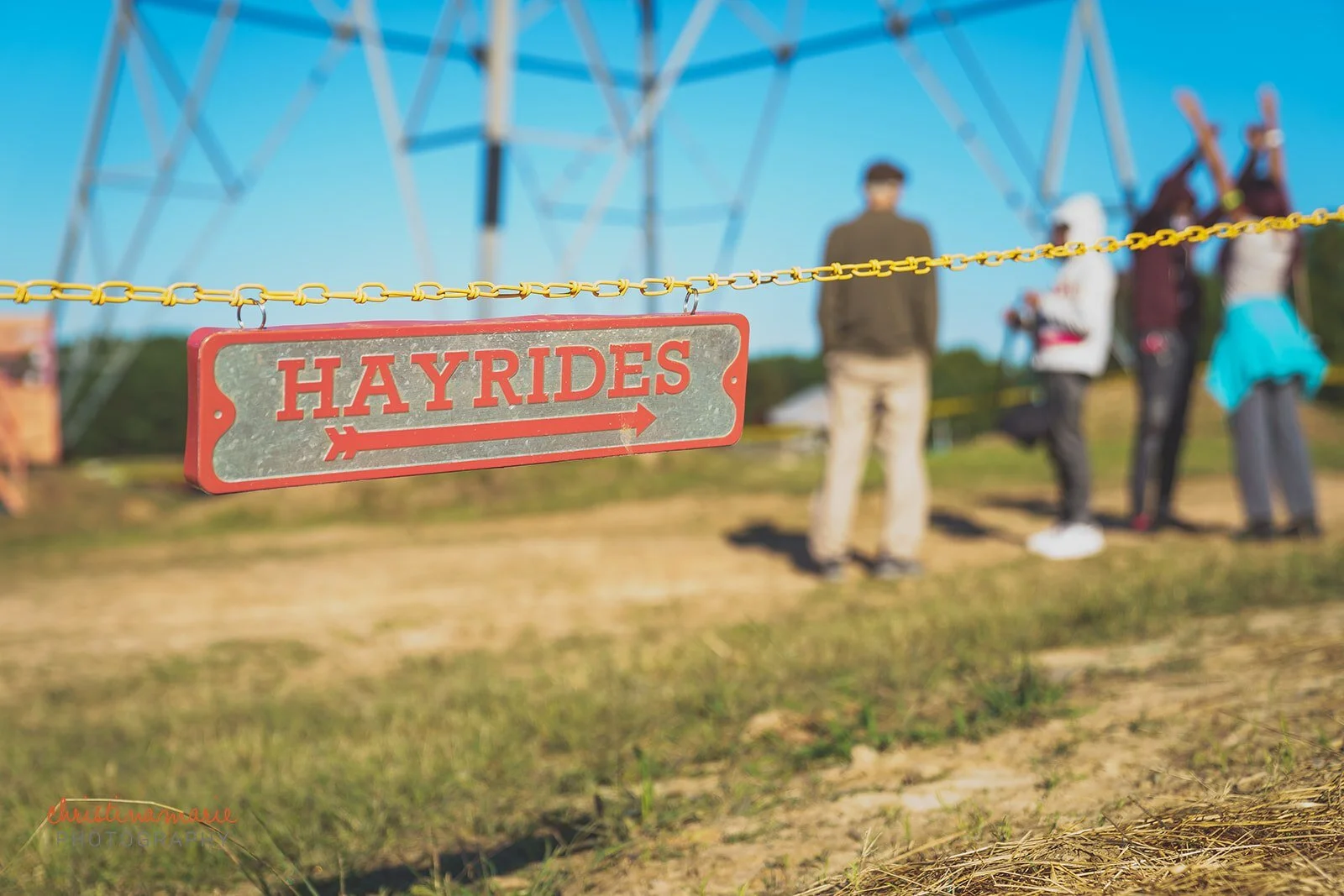 A red and gray sign hanging from a yellow chain with the word 'HAYRIDES' and an arrow pointing to the right. In the background, there are people standing near a large metal structure on a sunny day with a blue sky.