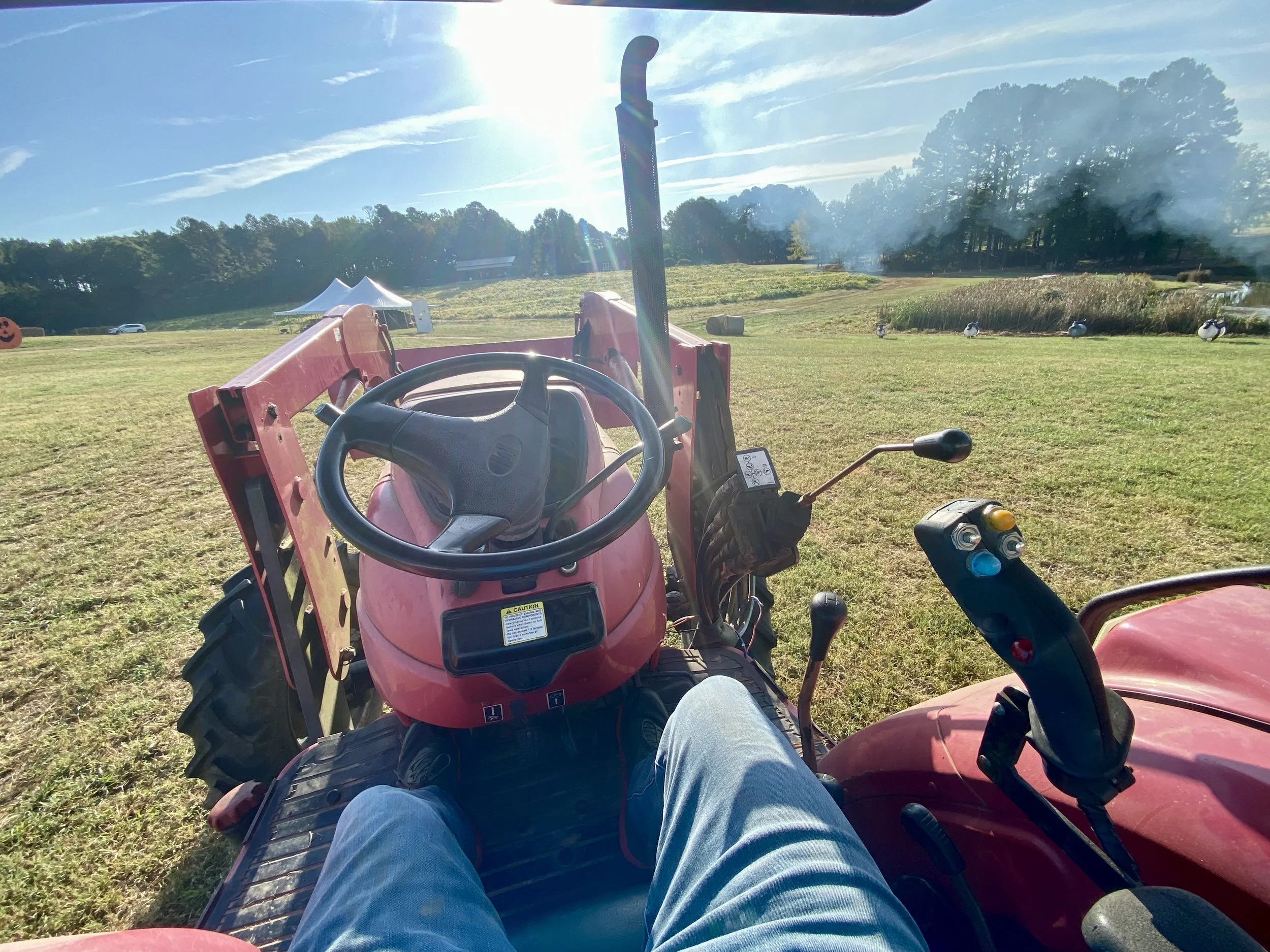 View from a person sitting on a small red tractor with a steering wheel and controls, looking out onto a grassy field with trees, tents, and a few ducks in the distance under a sunny sky.