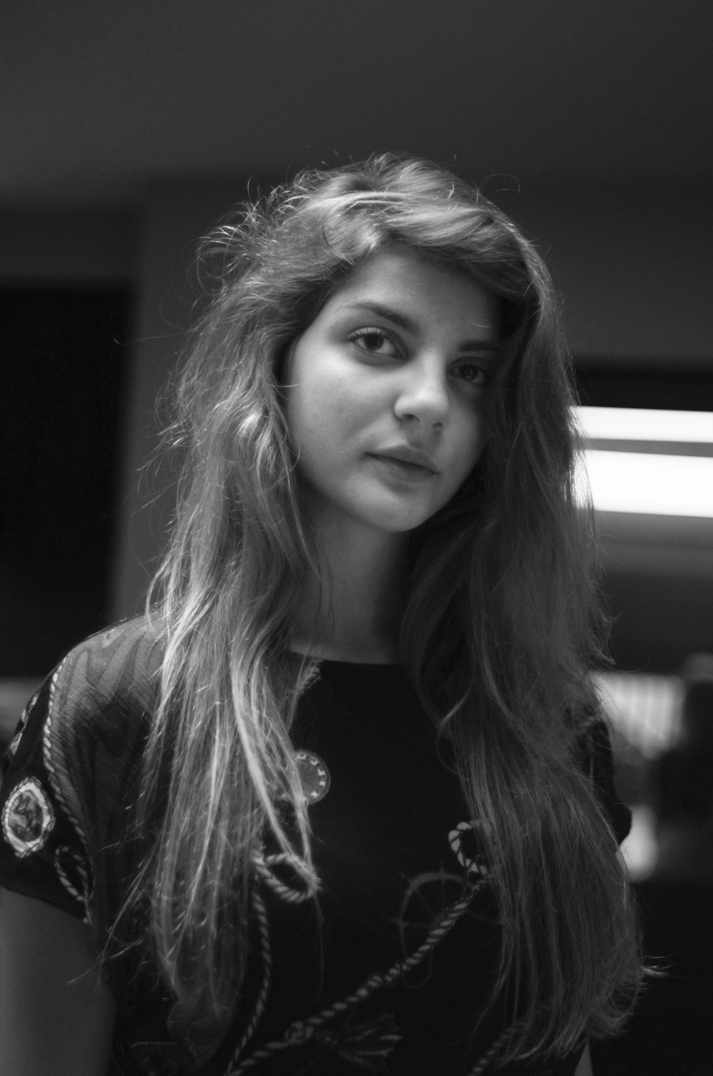 Black and white photo of a young woman with wavy hair, looking at the camera with a neutral expression. She is wearing a patterned top and appears to be indoors.
