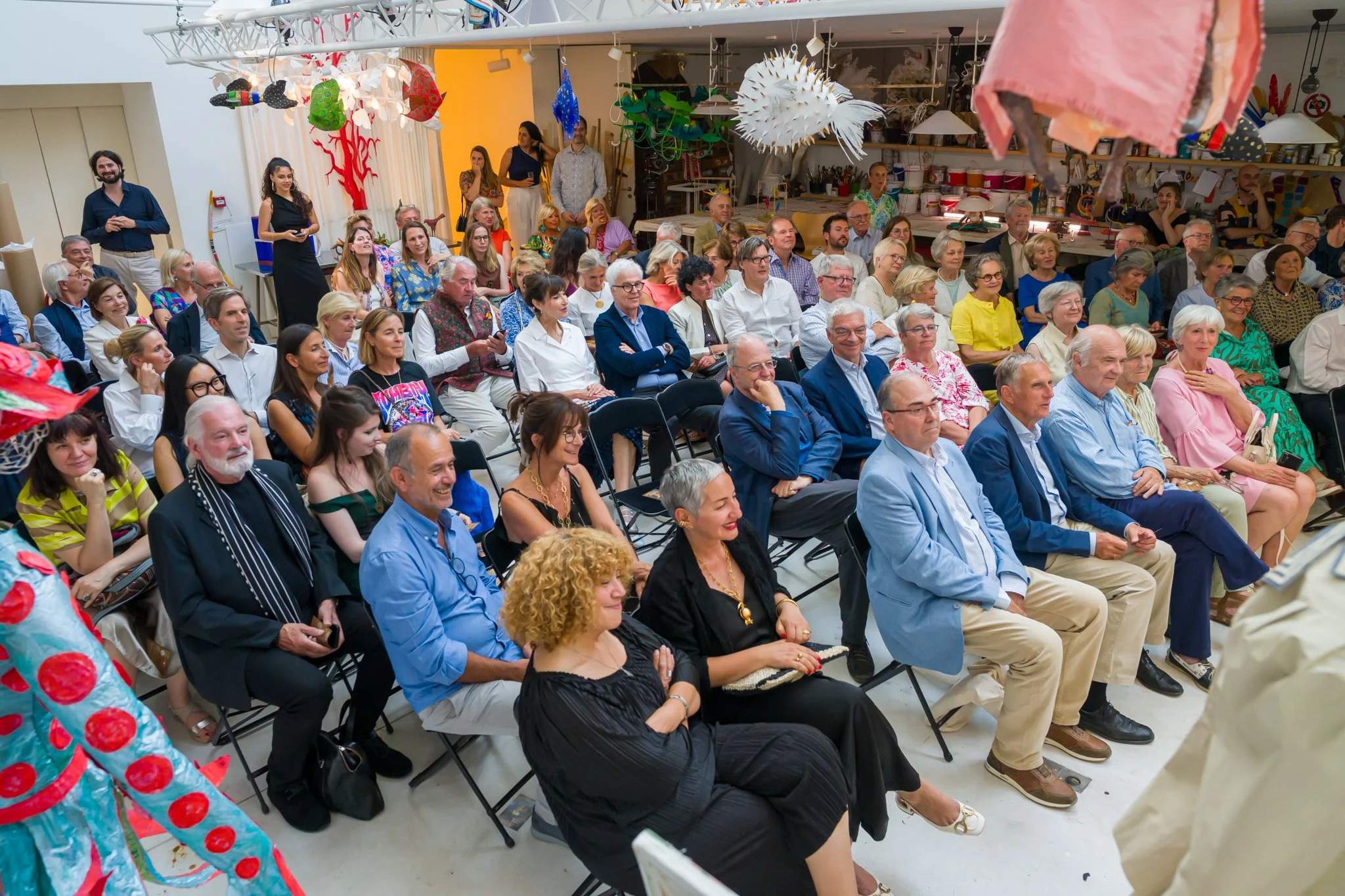 Un groupe de personnes assises dans une salle, attentives à une présentation ou un discours, avec des décorations colorées suspendues au plafond.