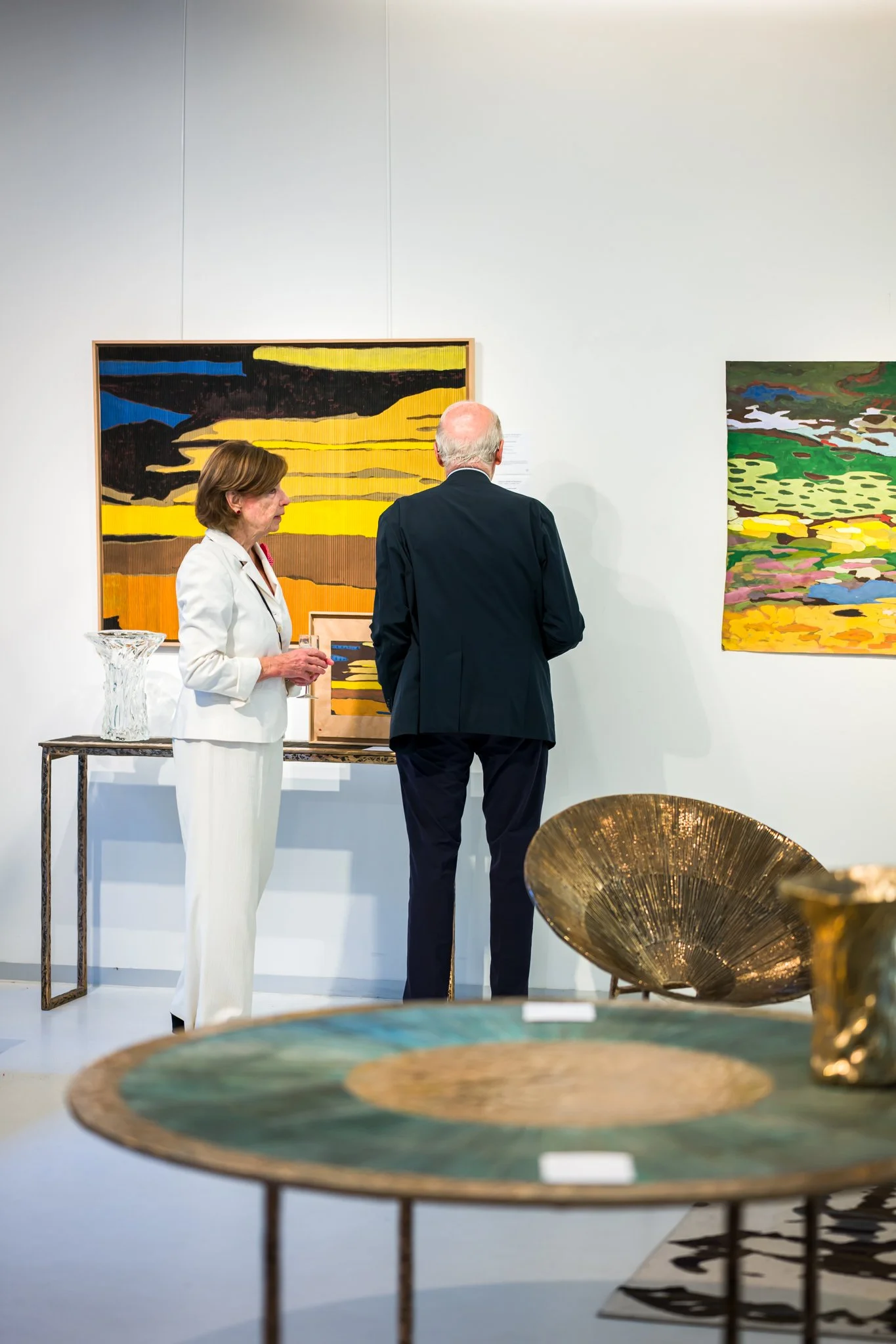 Deux personnes regardent des œuvres d'art dans une galerie, entourées d'objets décoratifs en or et d'une peinture abstraite colorée.