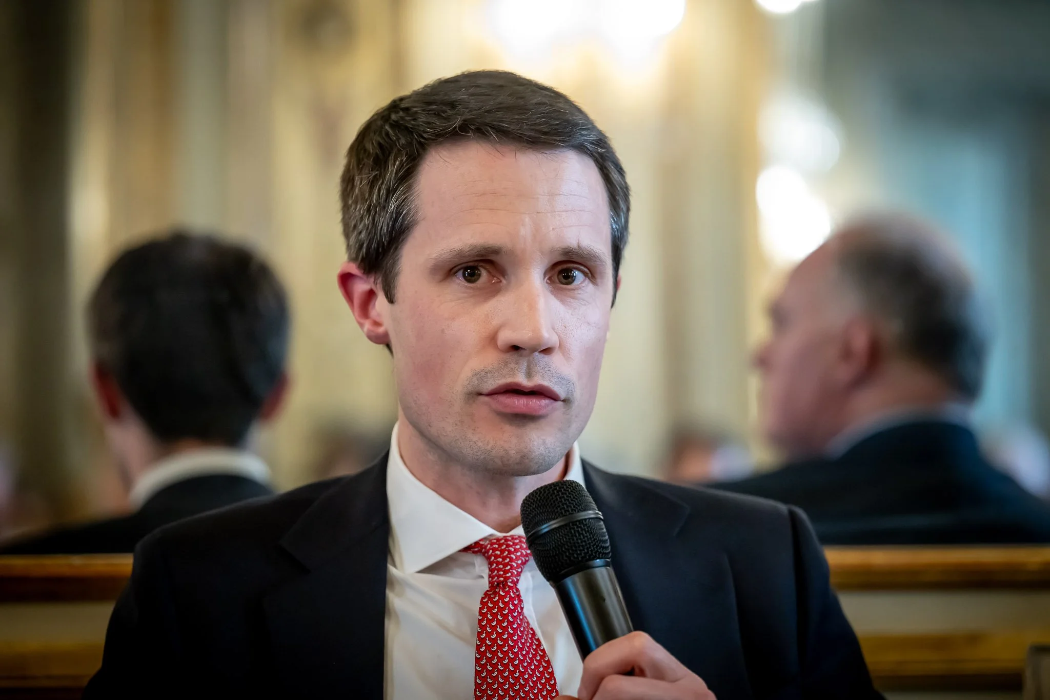 Man in a suit holding a microphone, speaking at an event.