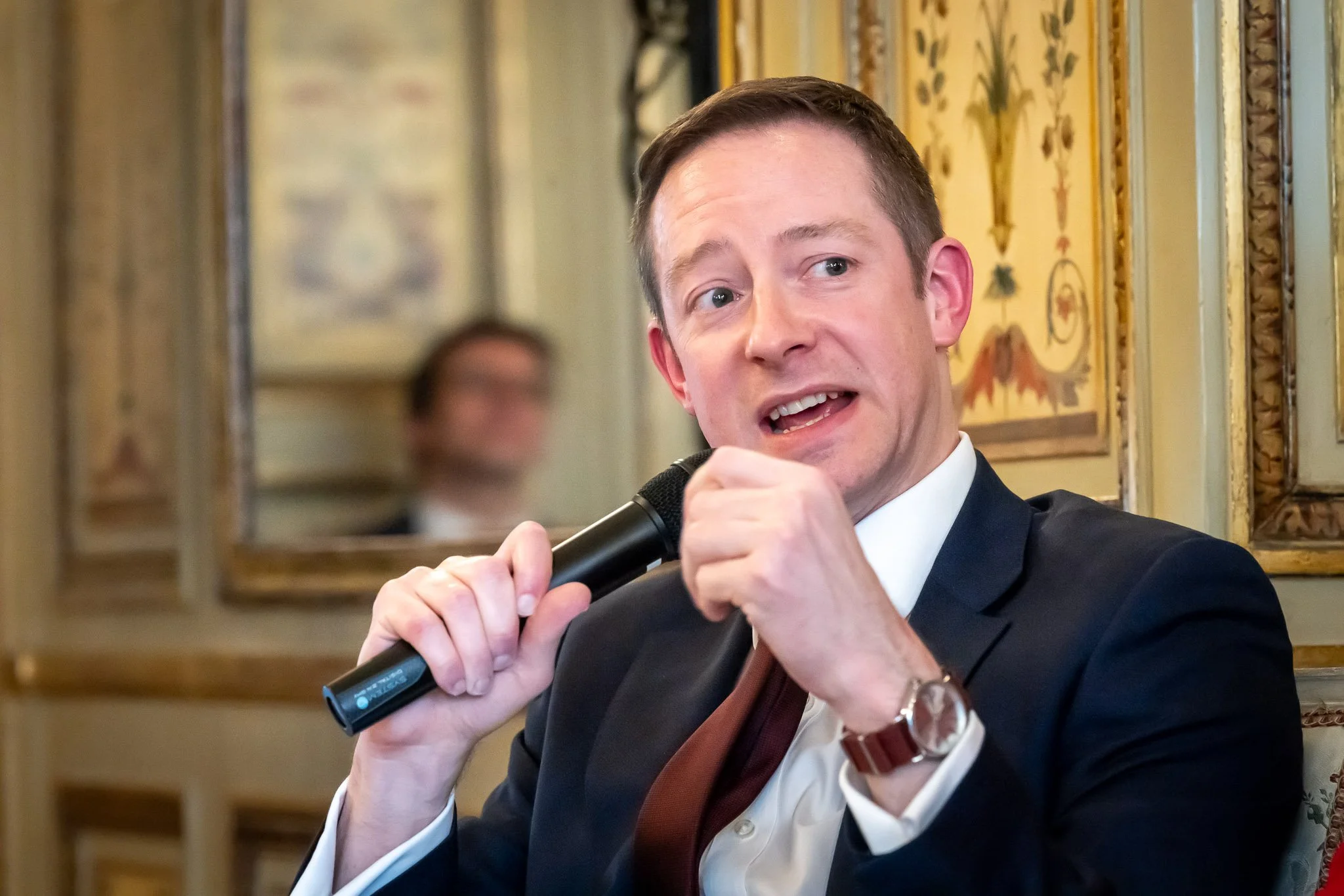 A man in a suit holding a microphone, speaking at an event or conference, with an elegant interior decor in the background.