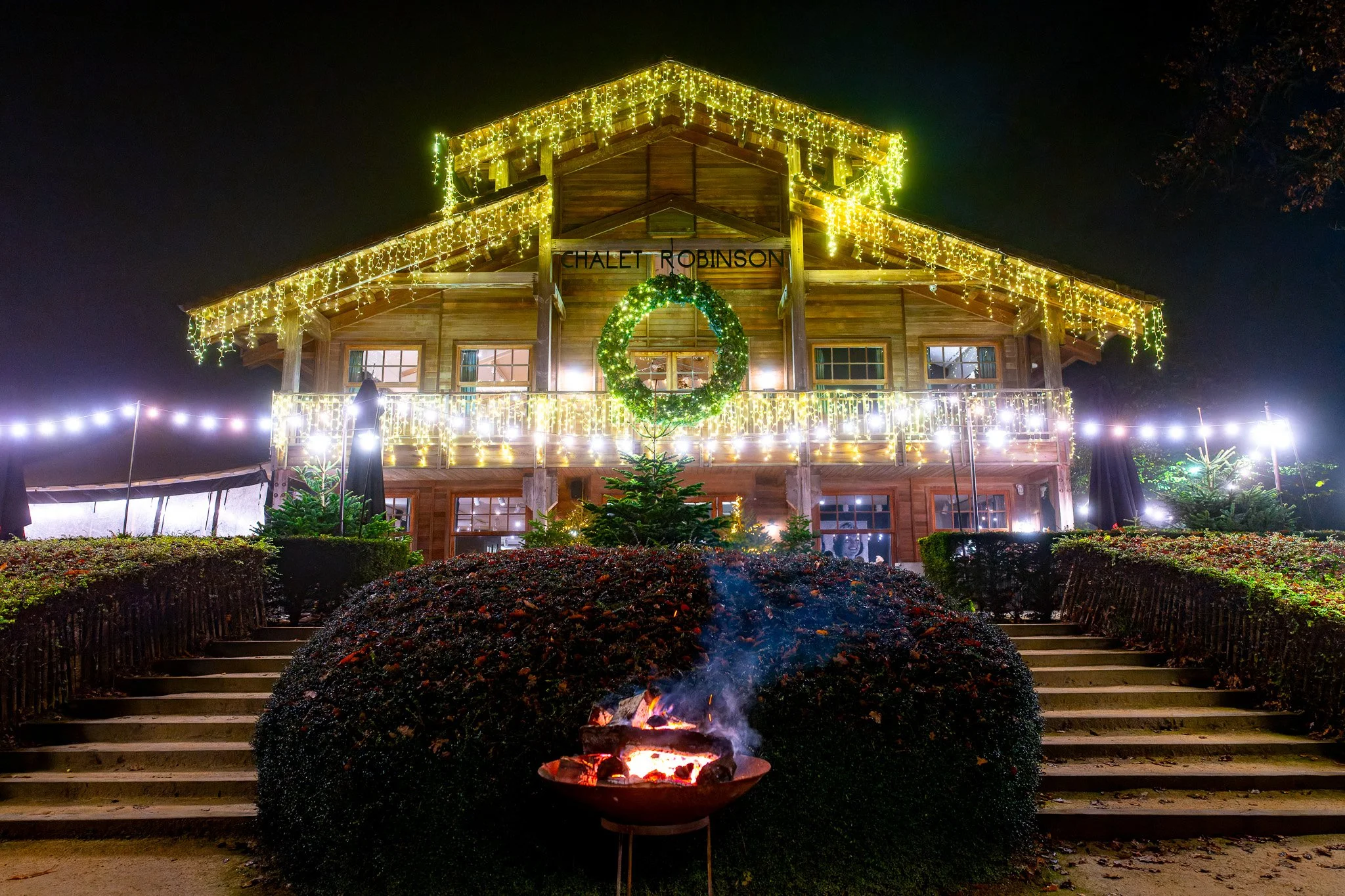 Wooden chalet decorated with Christmas lights at night, with a campfire in the foreground, surrounded by bushes and steps leading up to the building.
